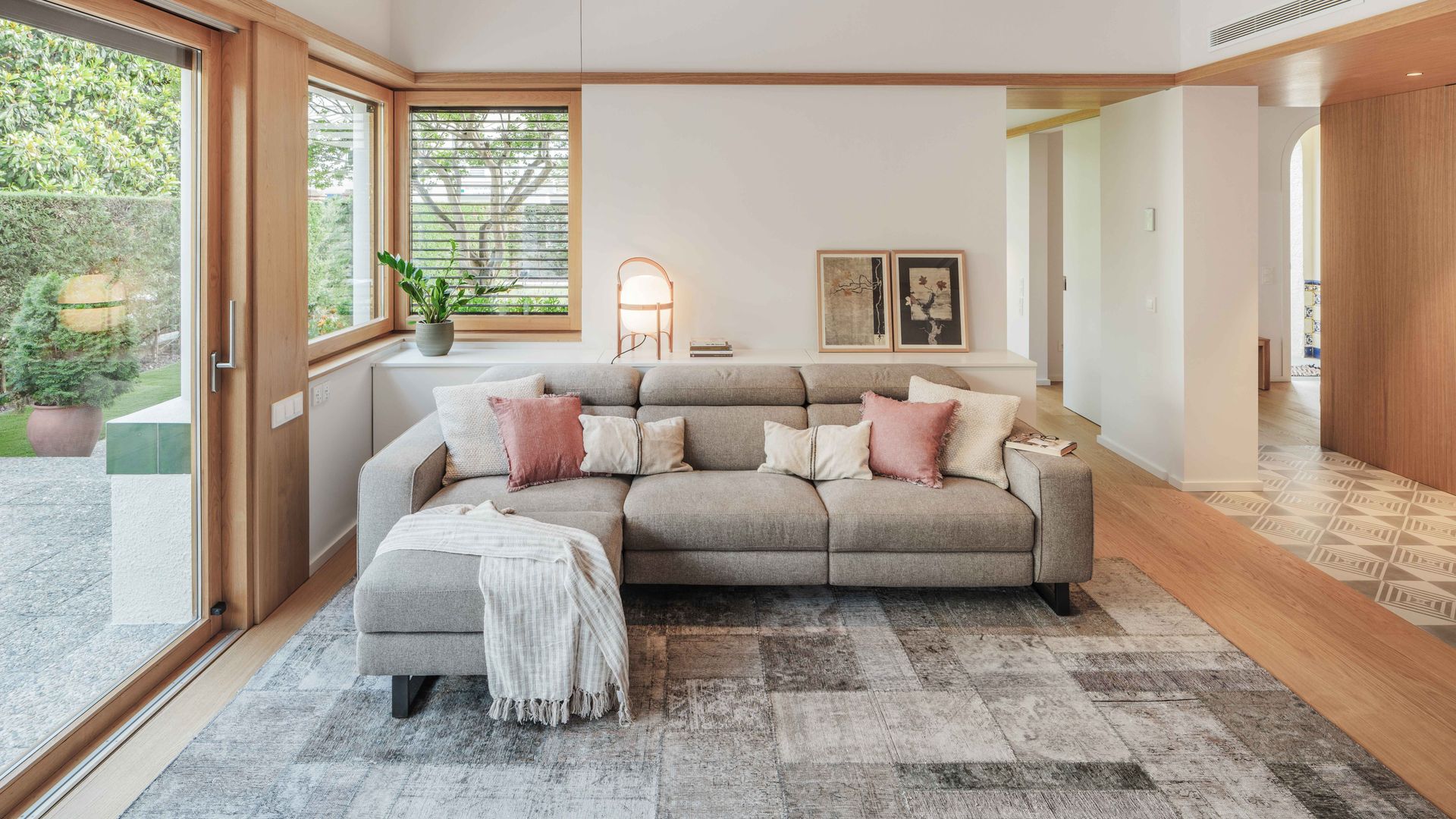 Salón con sofá gris con chaise longue, alfombra gris. aparador con lámpara Cesta, cuadros, puerta corredera de madera con salida al jardín