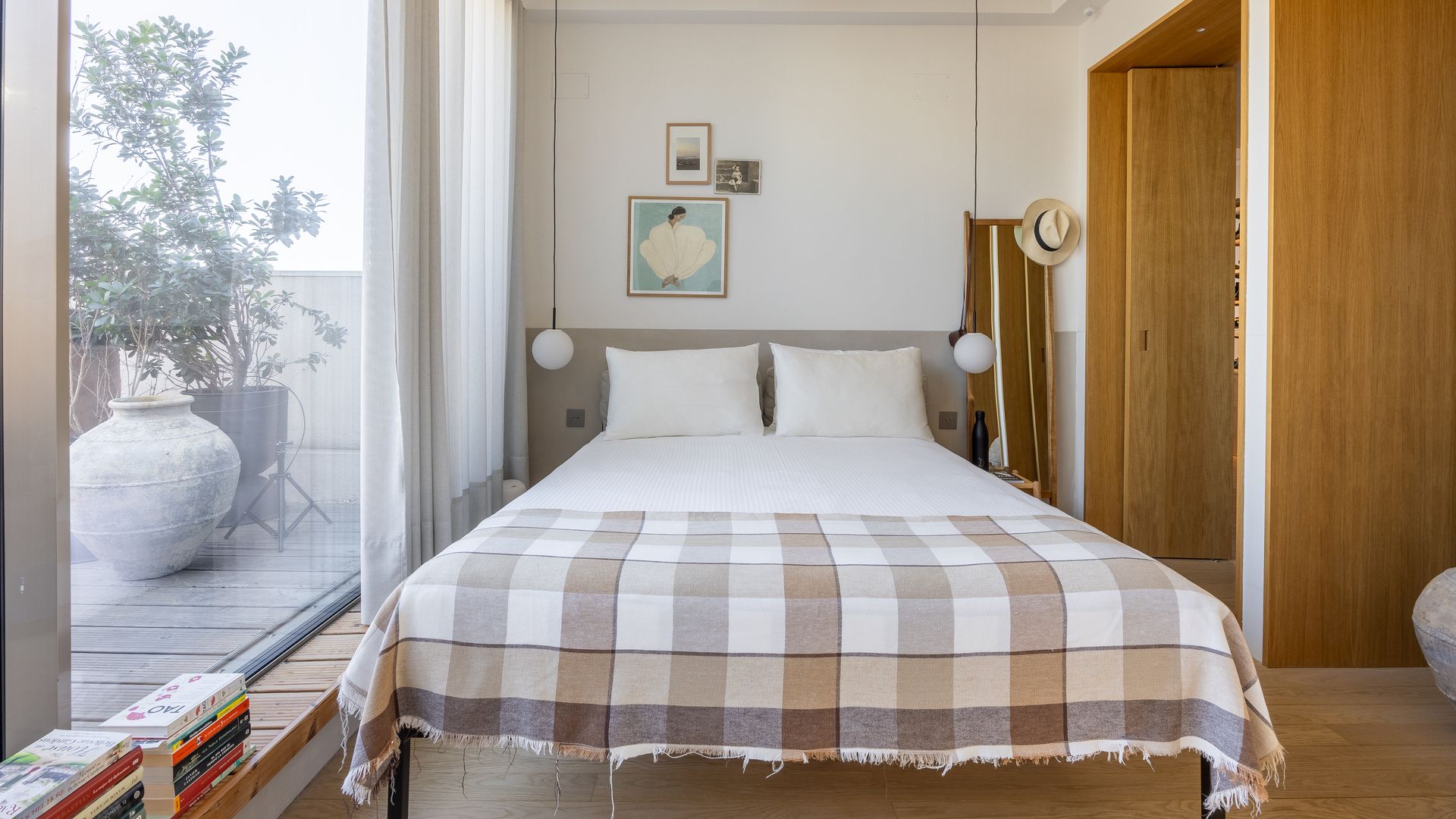 Dormitorio con la cama junto a la salida a la terraza, cabecero tapizado, manta de cuadros marrones, puertas de madera, lámparas de techo a ambos lados de la cama, libros en el suelo, cuadros en la pared del cabecero 