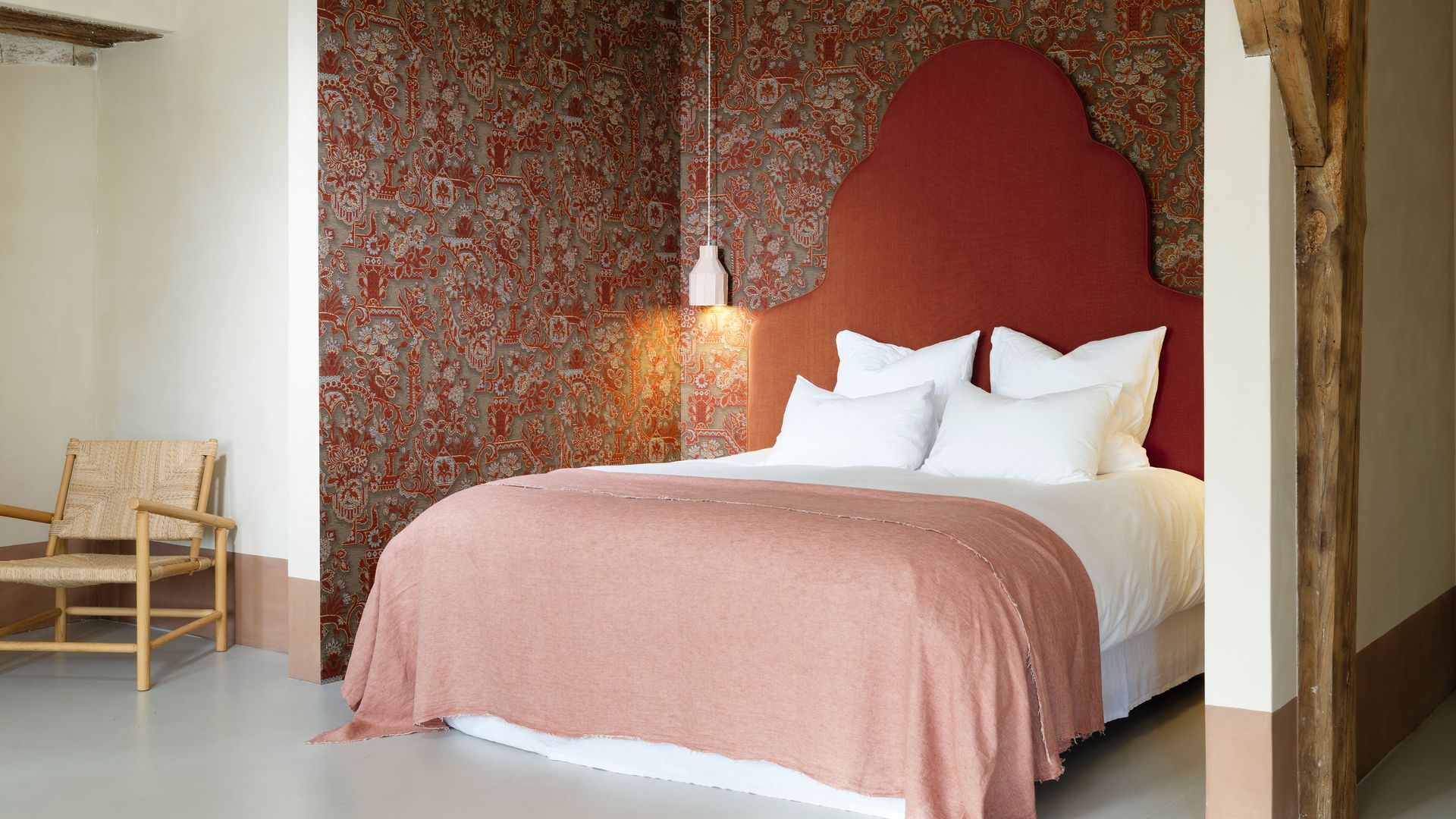 Dormitorio con cama con cabecero alto en forma de pico y color marrón, papel pintado en la pared del cabecero y en las de los lados, ropa de cama blanca, plaid caldero claro 