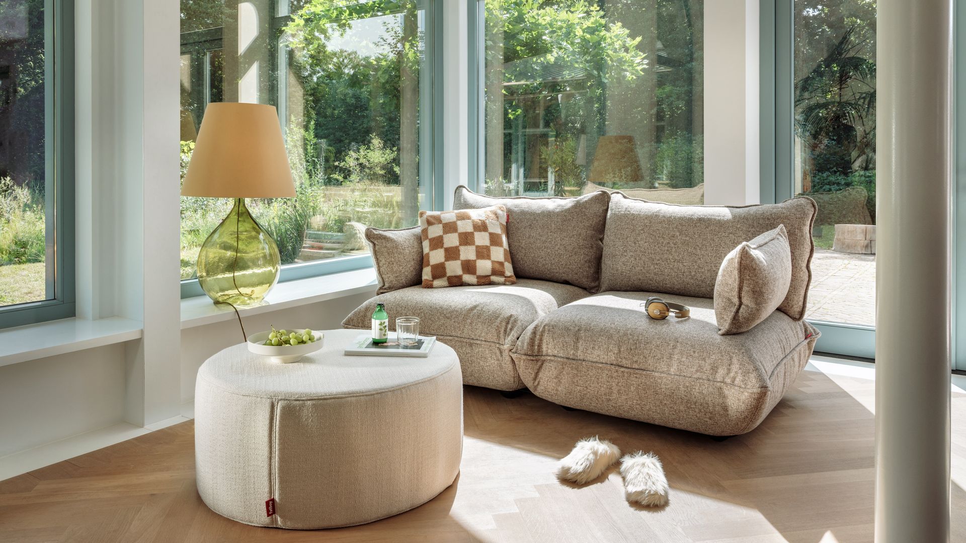 Salón con grandes ventanales con vistas a un jardín, alféizar ventana con banco y estantería, sofás mullido, mesa de centro tapizada, lámpara de sobremesa con el pie de cristal 