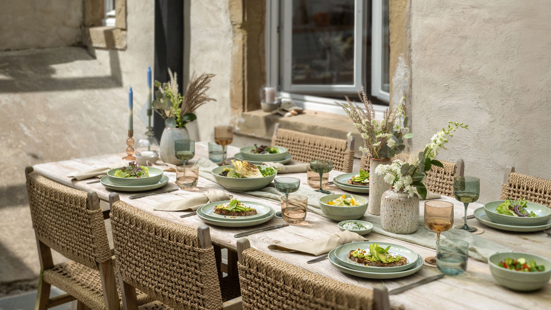 Mesa al aire libre con sillas de mimbre, vajilla de color verde, centros de flores y vasos y copas de colores 