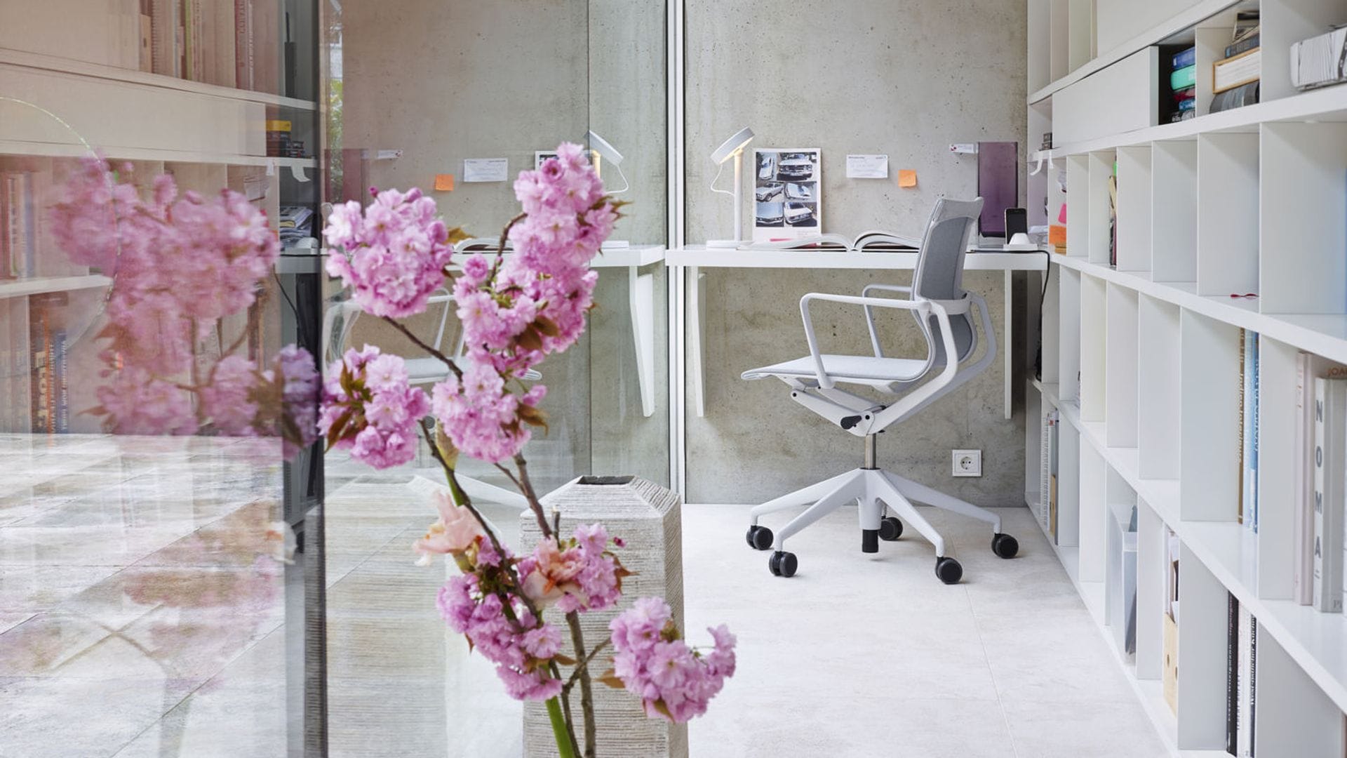 Despacho junto a gran ventanal con librería blanca, mesa blanca y silla con ruedas, orquídea rosa