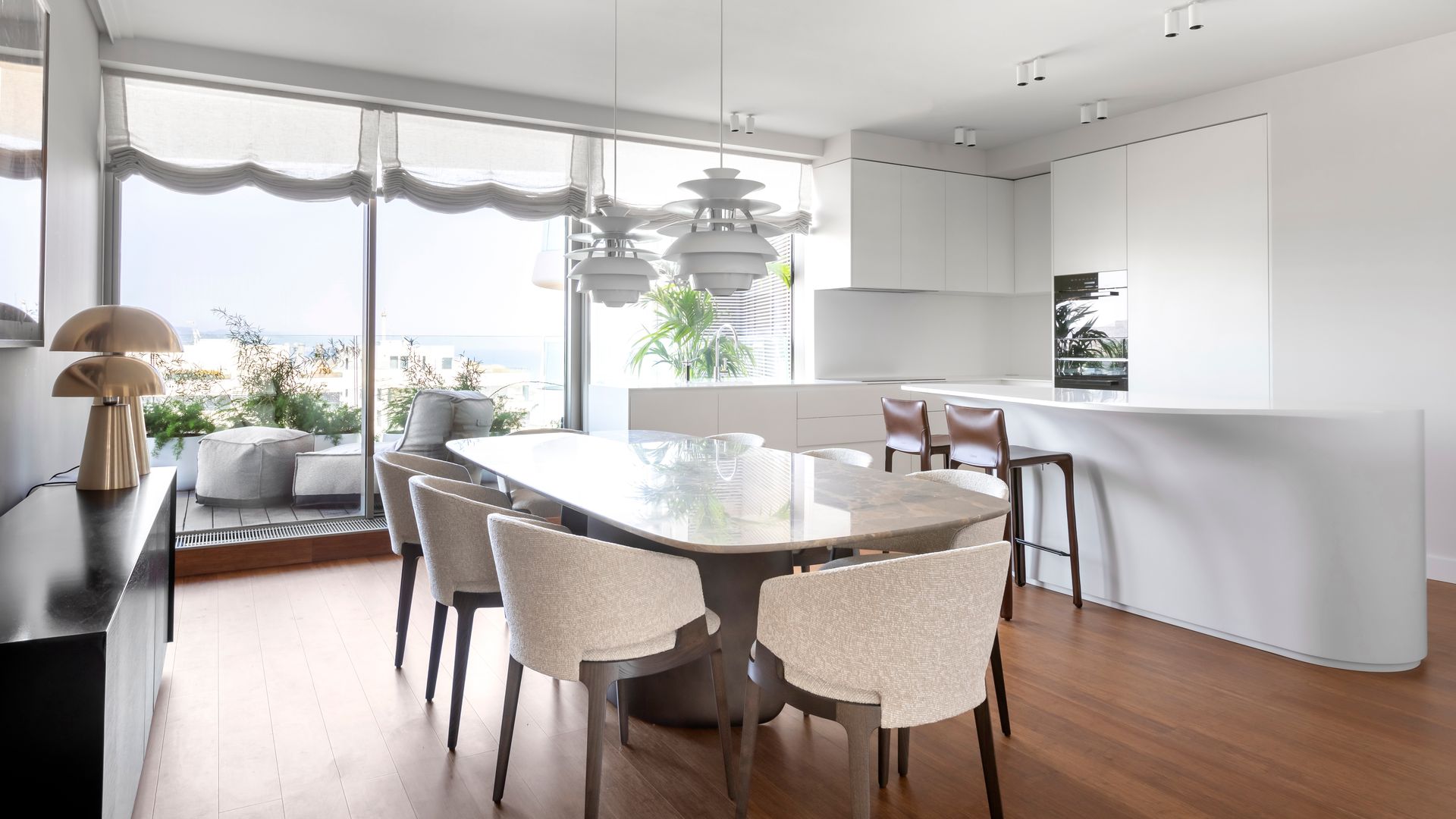 Cocina con comedor con salida a la terraza, estores blancos, mesa de comedor ovalada, sillas de comedor tapizada, cocina con isla, cocina blanca, suelo de madera, lámparas de techo 