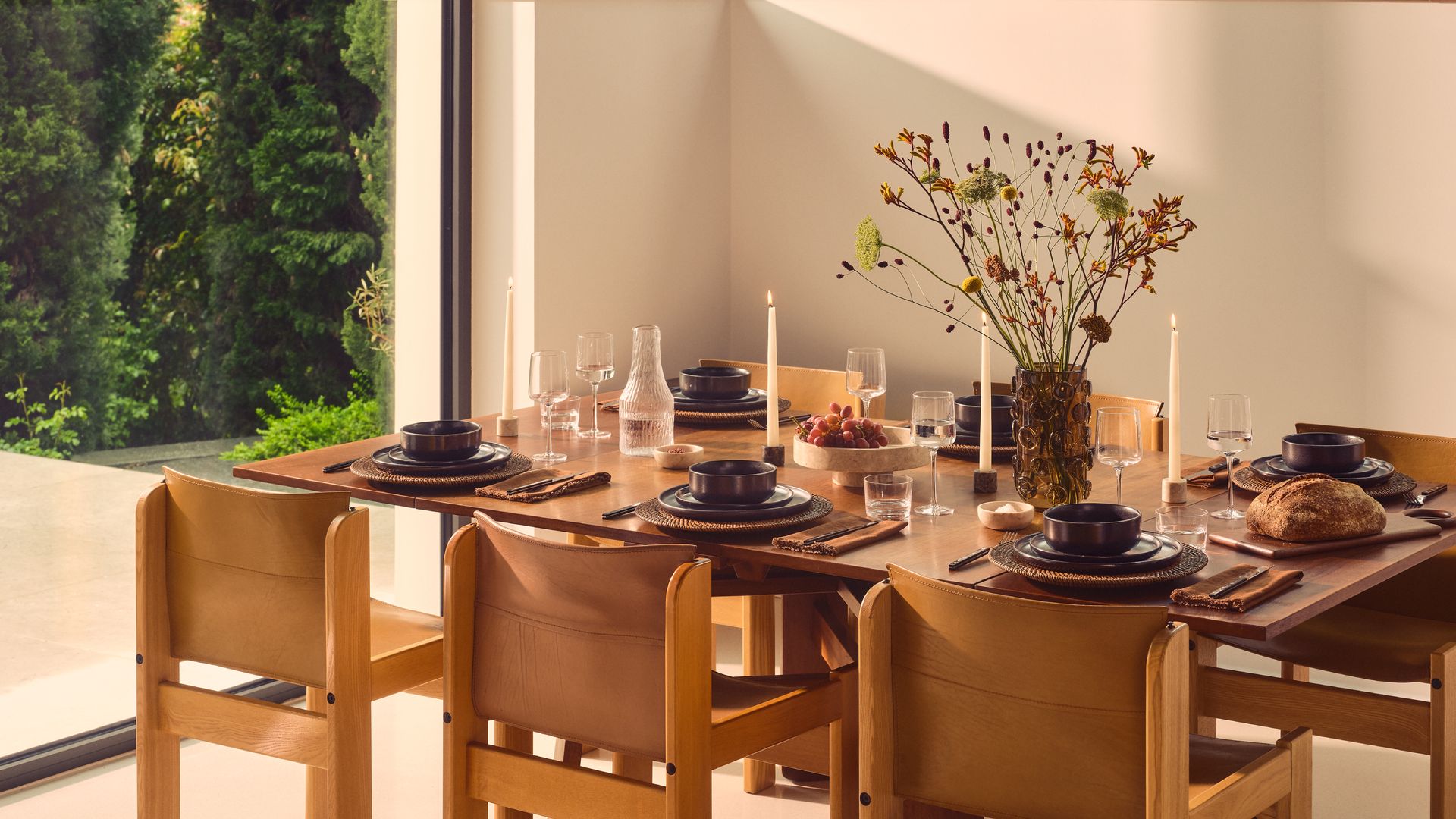 Comedor con mesa de madera, sillas de madera, florero, vajilla de color marrón oscuro, velas, botella de agua