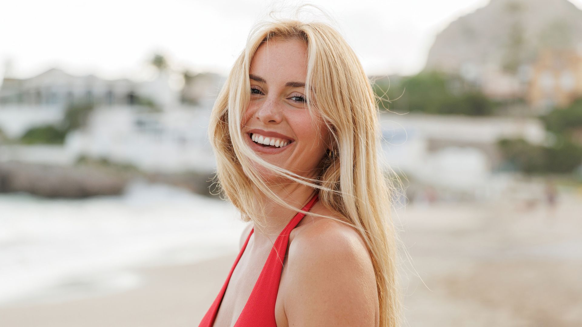 Mujer rubia con bañador rojo en la playa mirando sonriente a la cámara