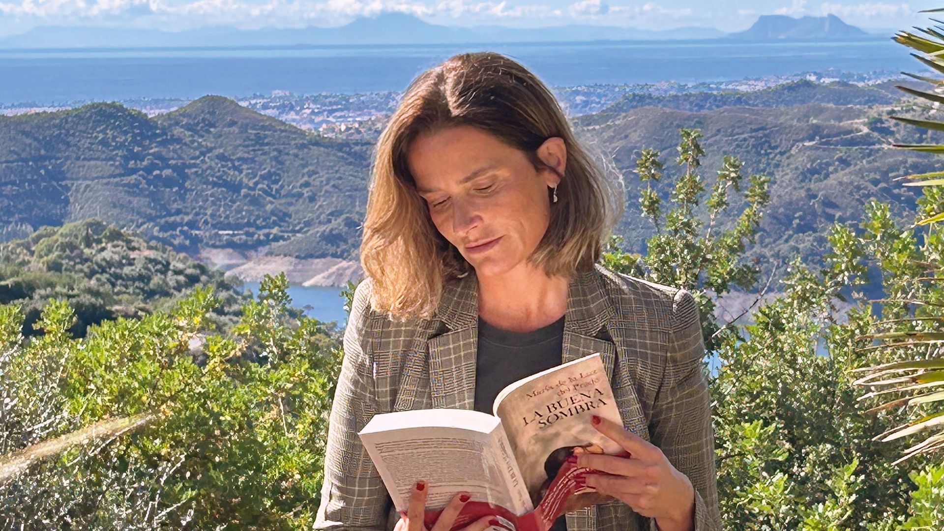 Mujer sentada en roca, leyendo libro en paisaje montañoso soleado.