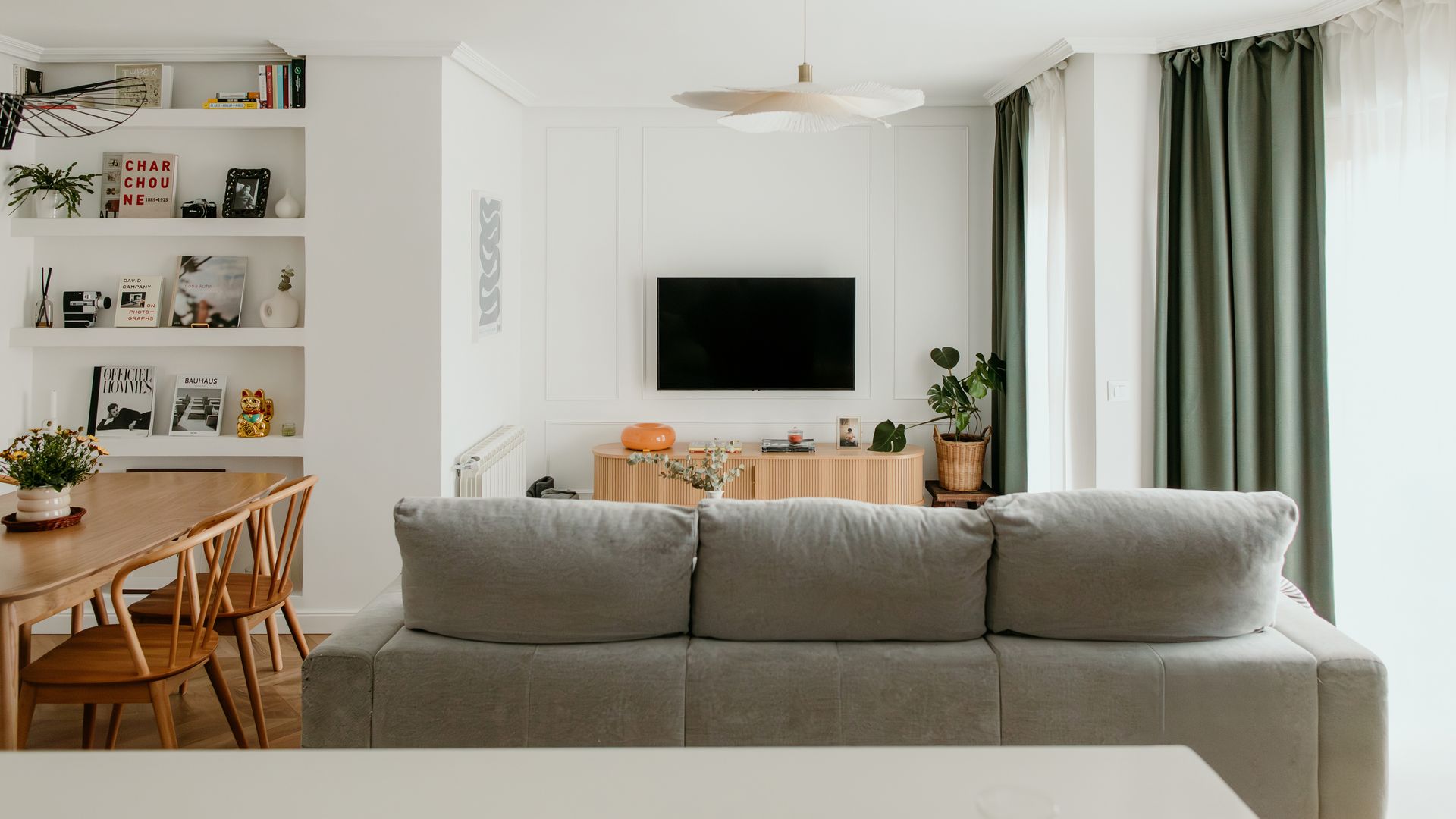 Salón comedor con molduras en la pared, televisor colgada de la pared, sofá gris, comedor con mesa y sillas de madera, lámpara de techo