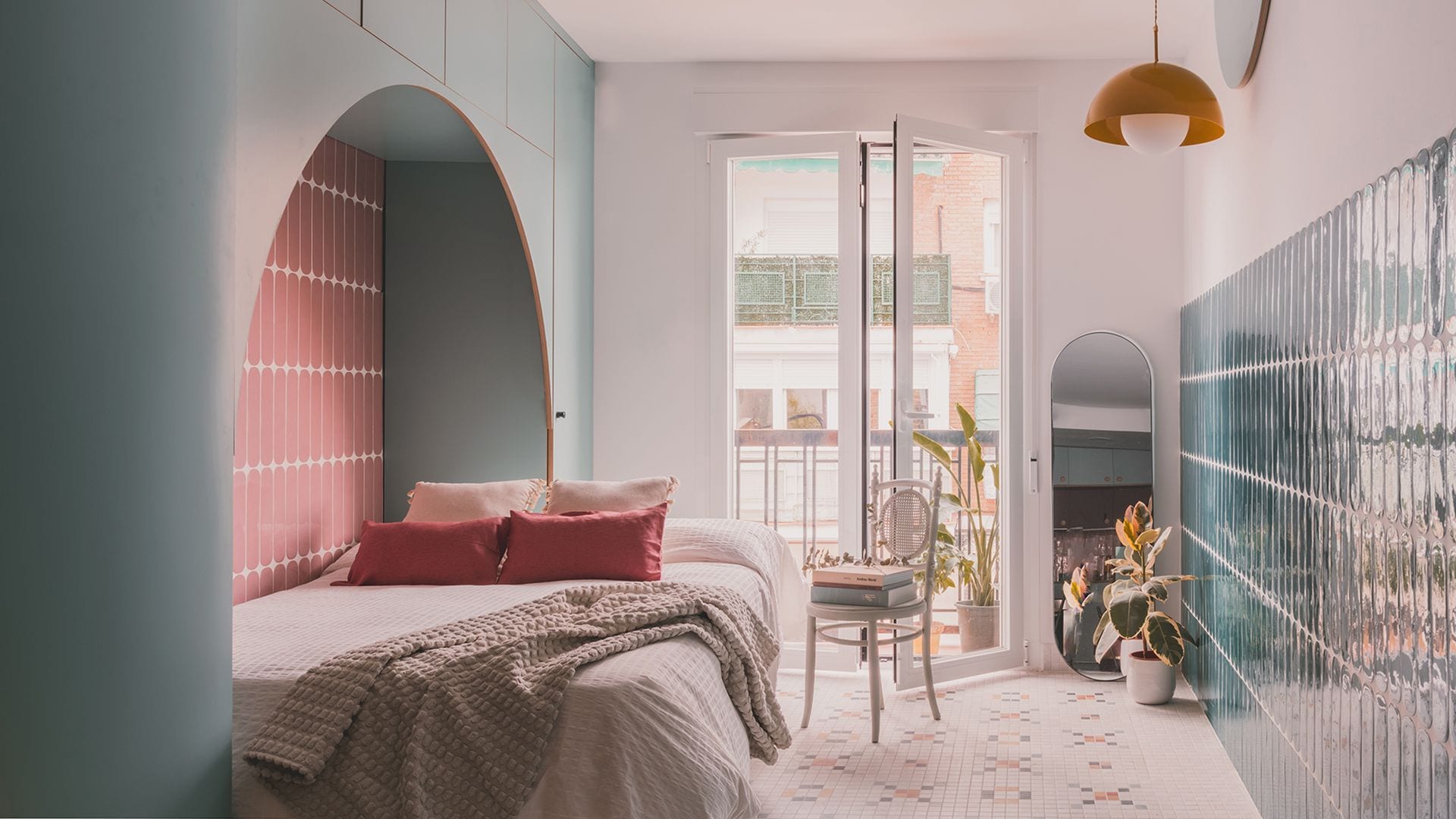 Dormitorio con salida a balcón, pared de azulejos azules, lámparas techo amarilla, cabecero a medida con azulejos rosas, silla, maceta con planta en el suelo, espejo de suelo, apoyado en la pared