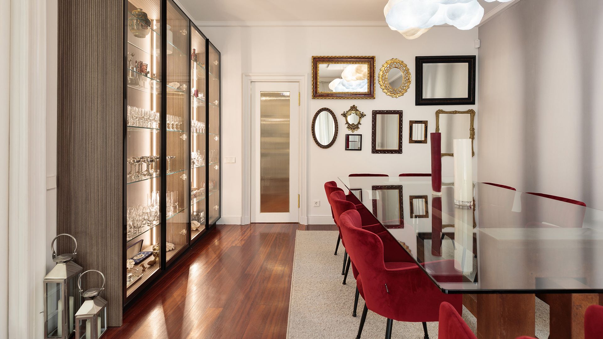 Comedor con mesa de cristal, vitrina de gran tamaño con vajilla y cristalería, cuadros y marcos en la pared, sillas rojas, alfombra