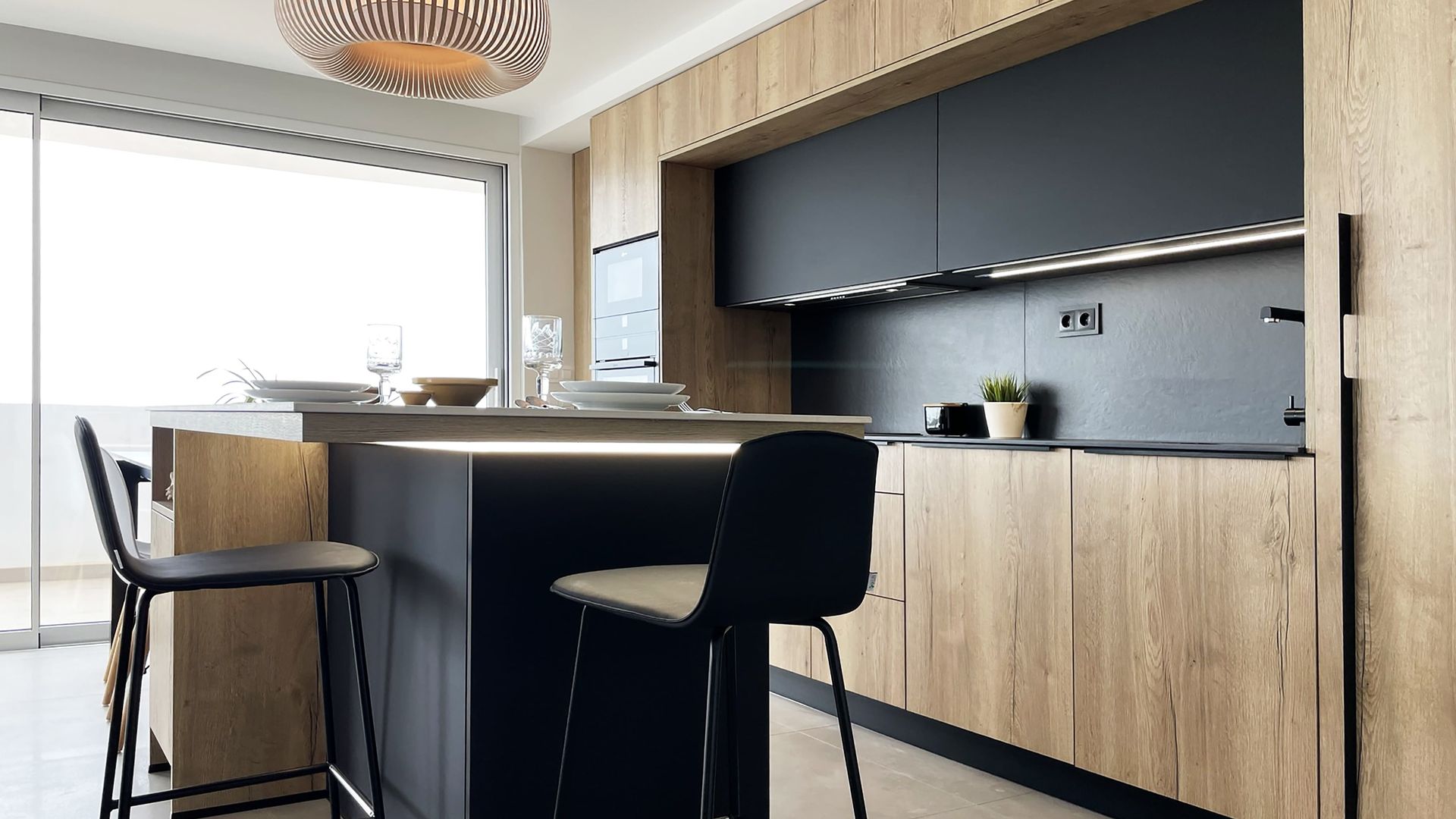 Cocina con isla con muebles de madera y en color negro, zona de barra con taburetes, luz bajo la encimera de la isla, lámpara de techo