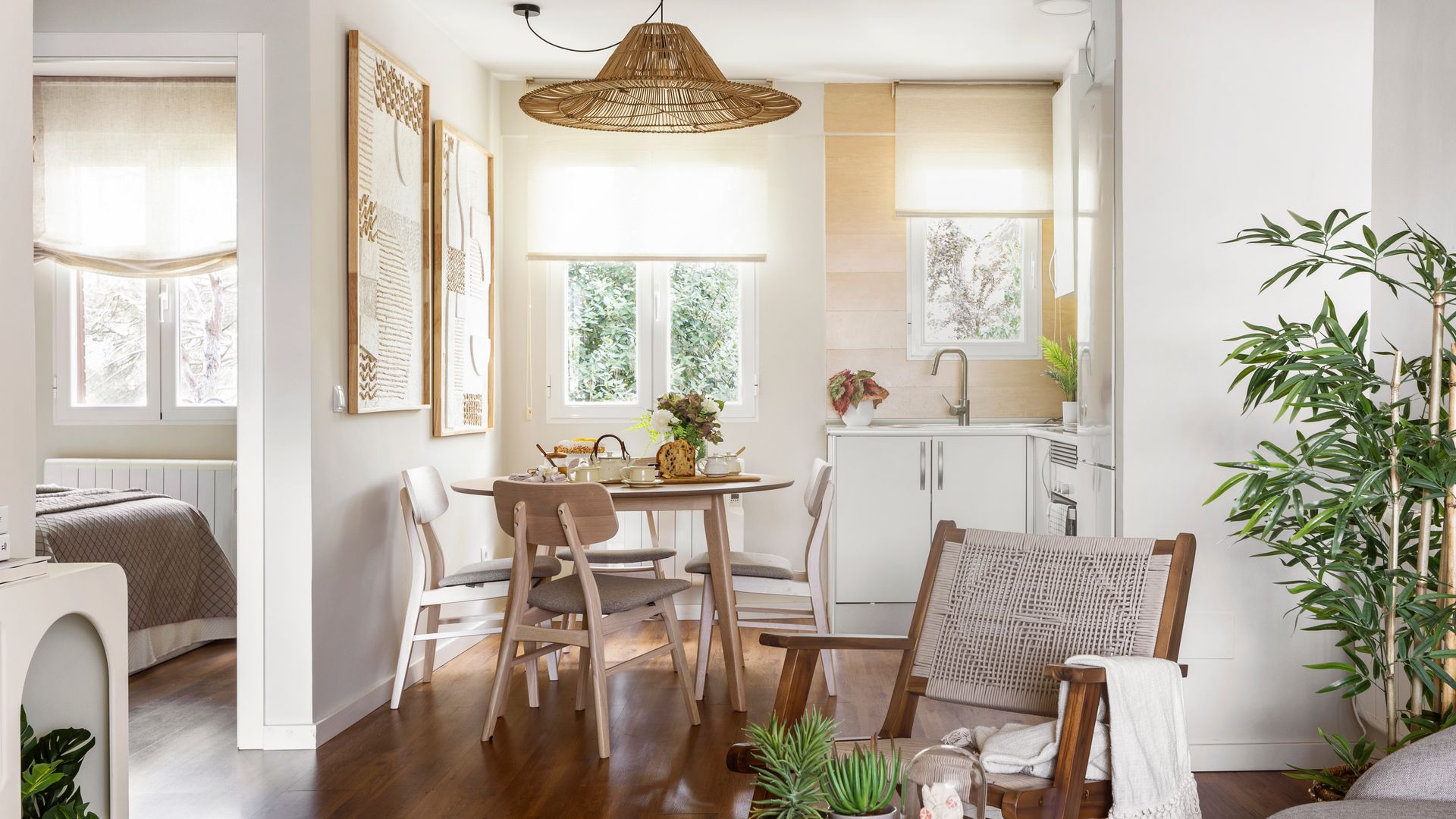 Cocina abierta al salón con zona de office, suelo de madera oscura, muebles blancos, sofá gris, cojines, lámpara de techo de fibras vegetales, sillas de madera 