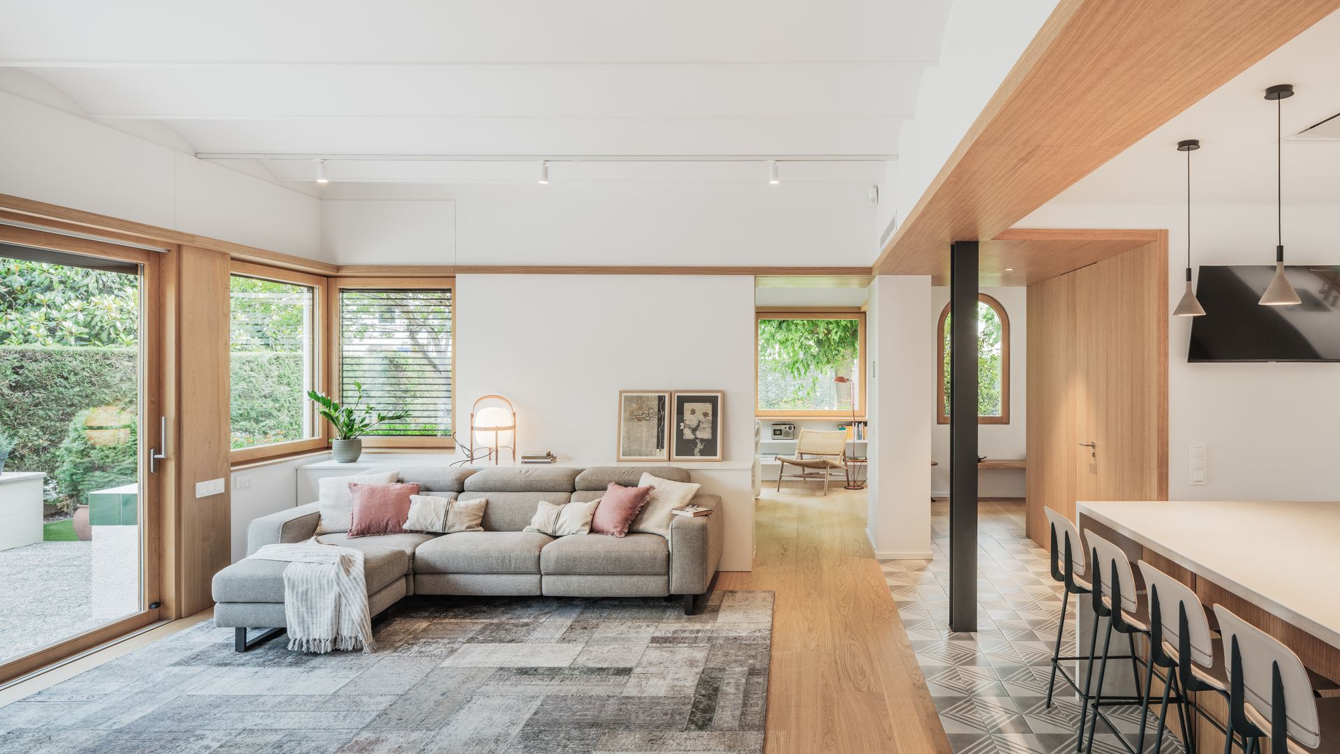Salón abierto a la cocina, la entrada y el comedor, con una gran corredera de madera con salida al jardín, alfombra gris, sofá gris con chaise longue, cuadros sobre un mueble, lámpara Cesta, suelo de madera, suelo hidráulico, regleta de focos blancos en el techo