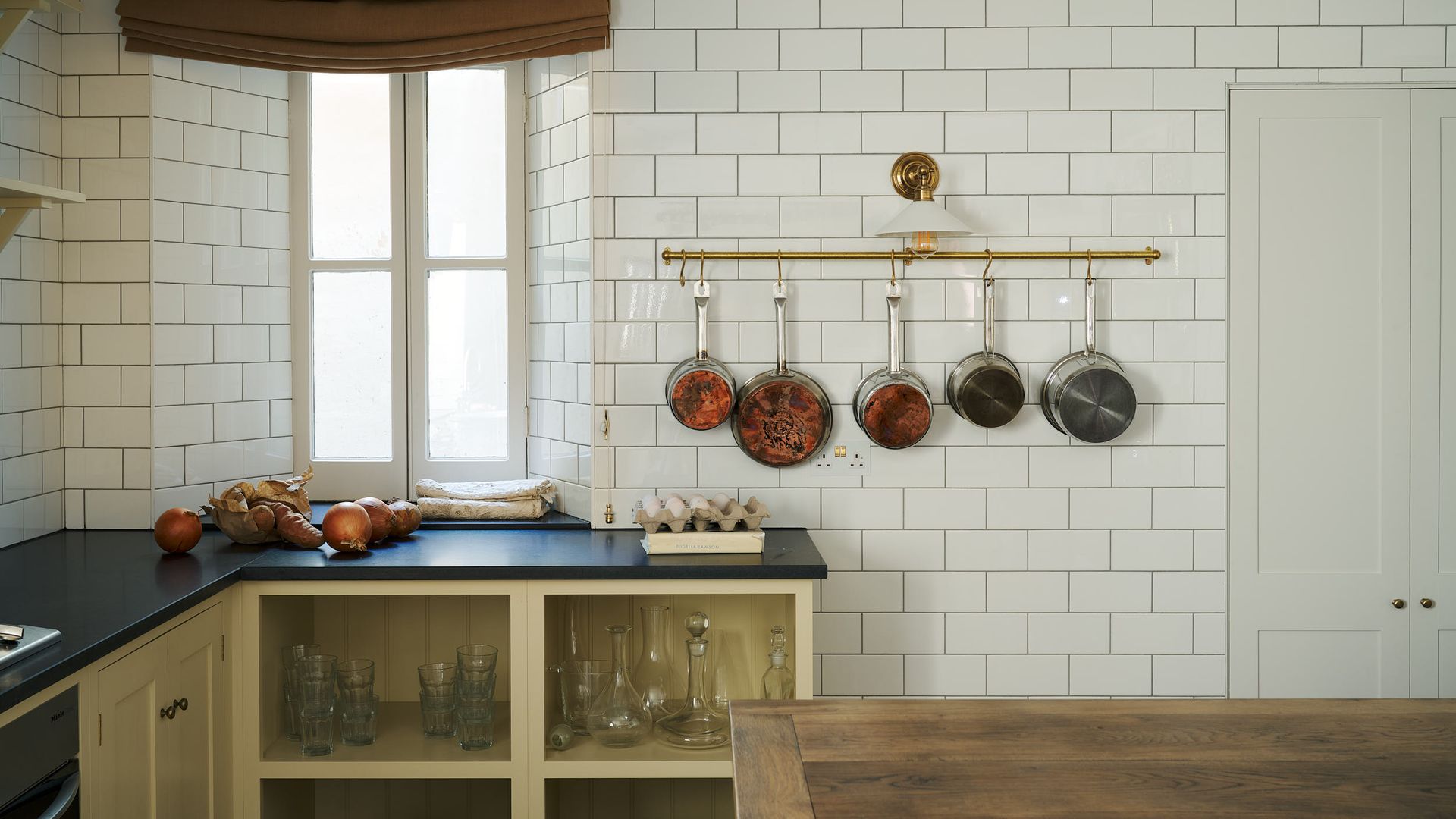 Cocina con pared de azulejos, barra dorada con sartenes, ventana con estor, encimera negra, muebles interiores abiertos con cristalería