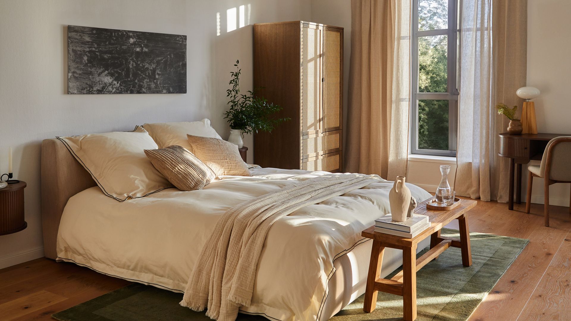 Dormitorio de verano con banco de madera a los pies de la cama, alfombra verde, armario de rejilla, cabecero tapizado, ropa de cama en tonos beiges y escritorio en madera