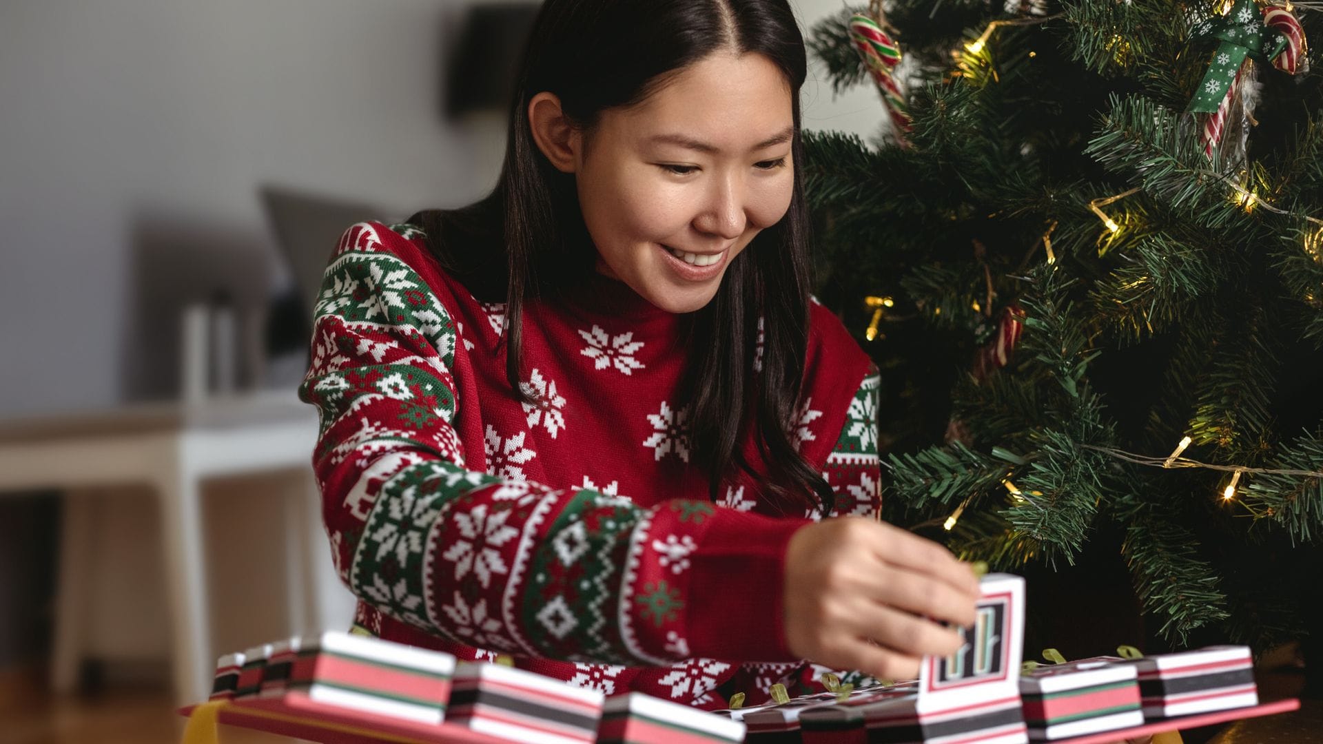 Mujer joven asiática con jersey navideño abriendo calendario de Adviento junto al árbol de Navidad