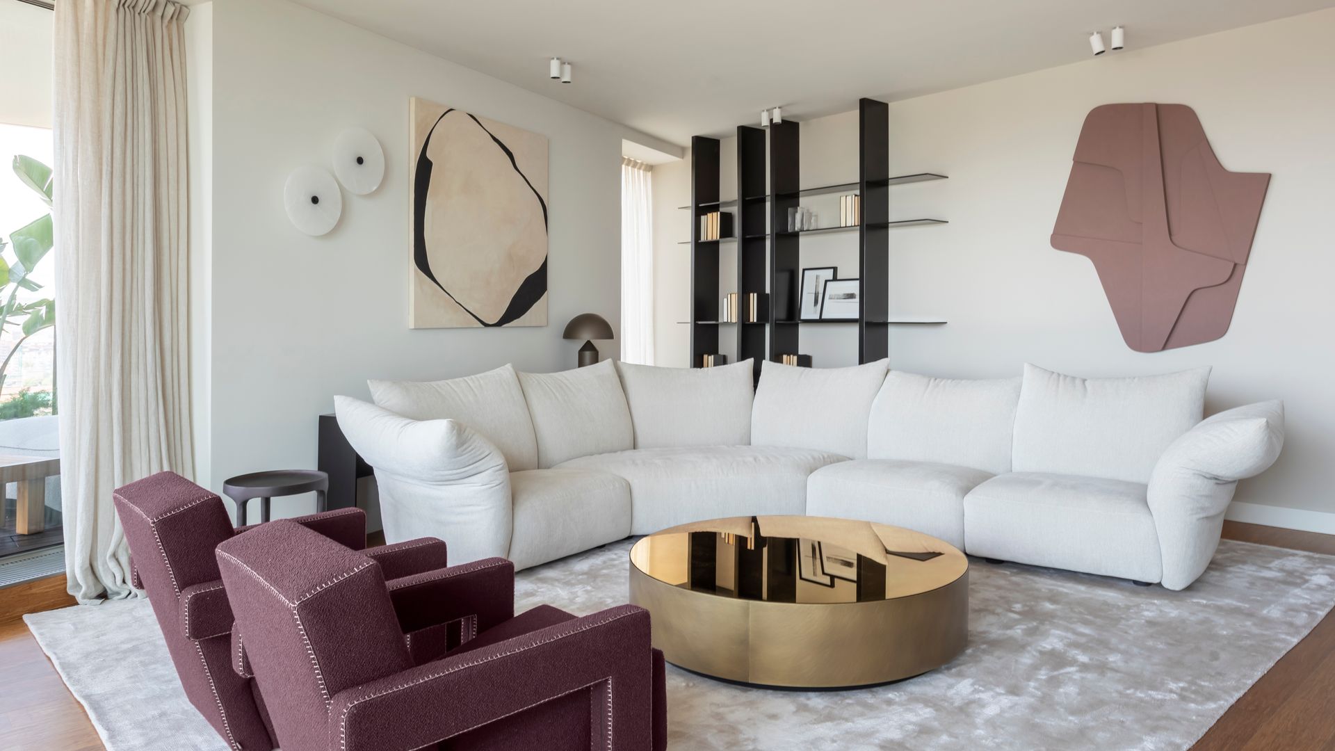 Salón con sofá blanco, butacas burdeos, mesa de centro dorada, alfombra, cortinas blancas y cuadros en la pared 