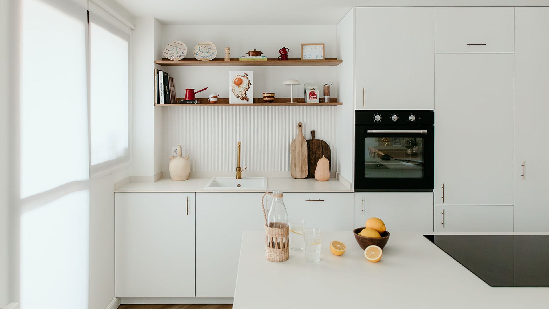 Cocina blanca con isla, baldas de madera, estores blancos, suelo de madera, horno negro