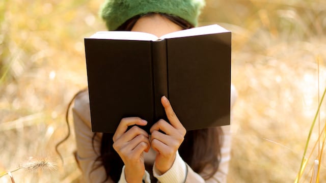 Mujer leyendo un libro de cubierta negra con gabardina y boina en el campo en otoño