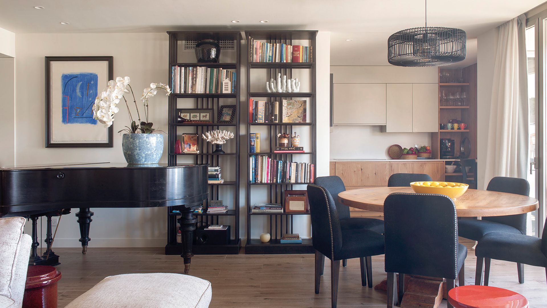 Salón comedor y cocina en uno con piano, mesa de comedor redonda de madera, sillas negras tapizadas y sofá beige