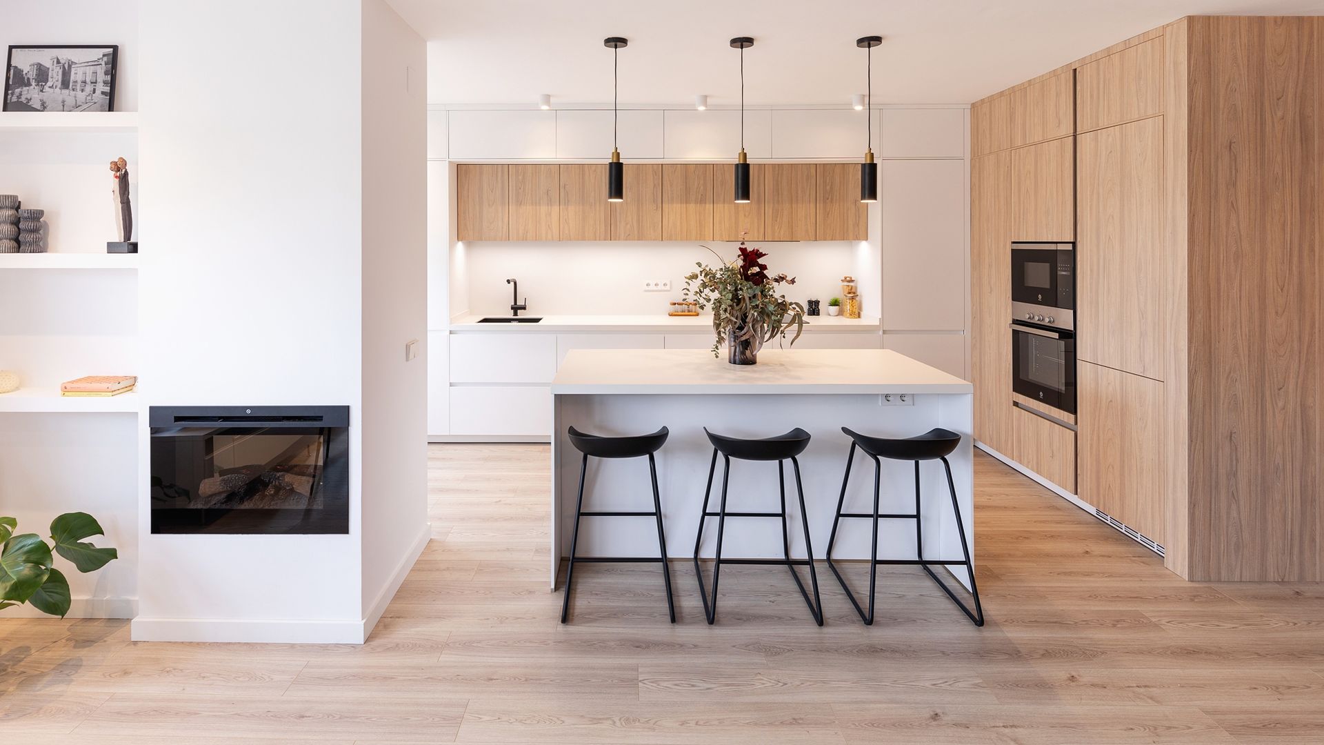 Cocina abierta con isla en blanco y madera, lámparas de techo sobre la isla, taburetes negros