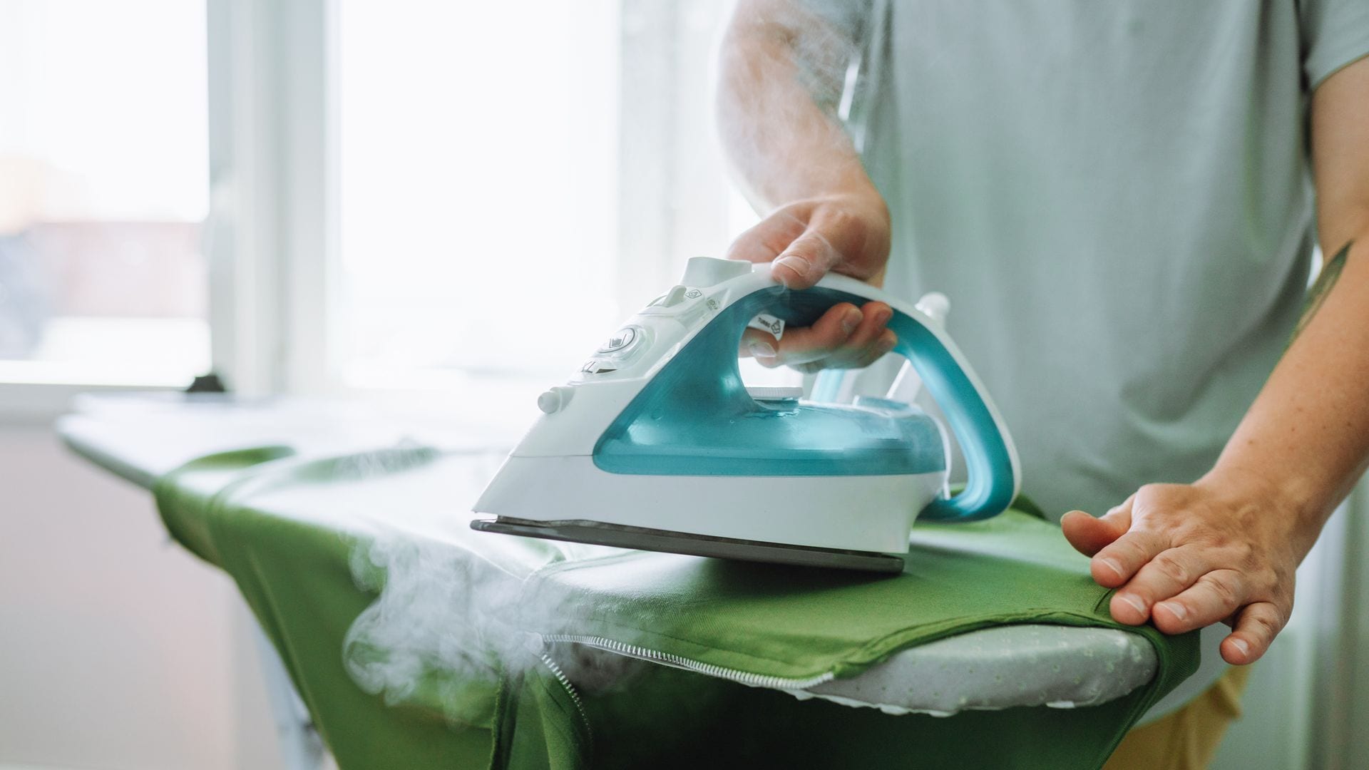 Hombre planchando con una plancha de vapor blanca y turquesa, sobre una tabla, una prenda verde.