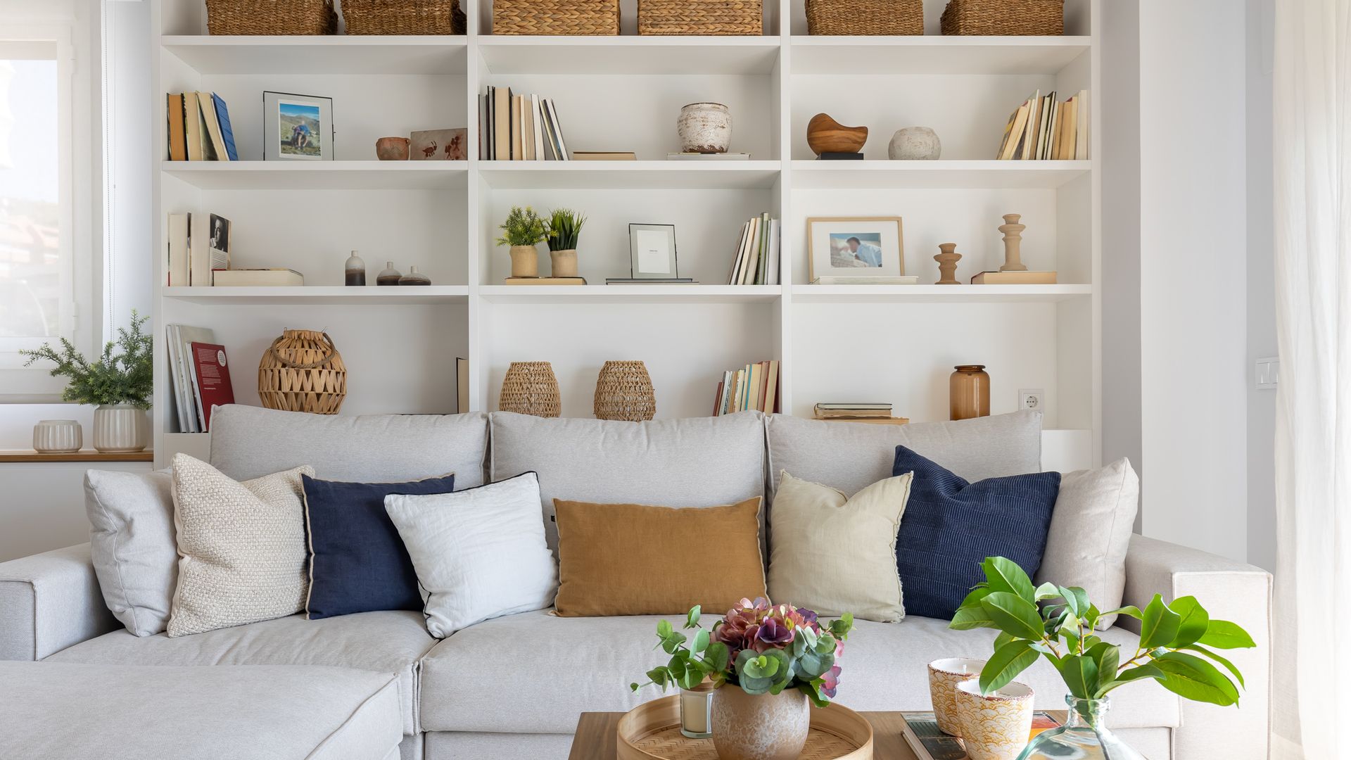 Salón con estantería blanca con fotos, cestos, libros y adornos, sofá blanco, mesa de centro de madera, adornos, plantas y bandejas sobre la mesa