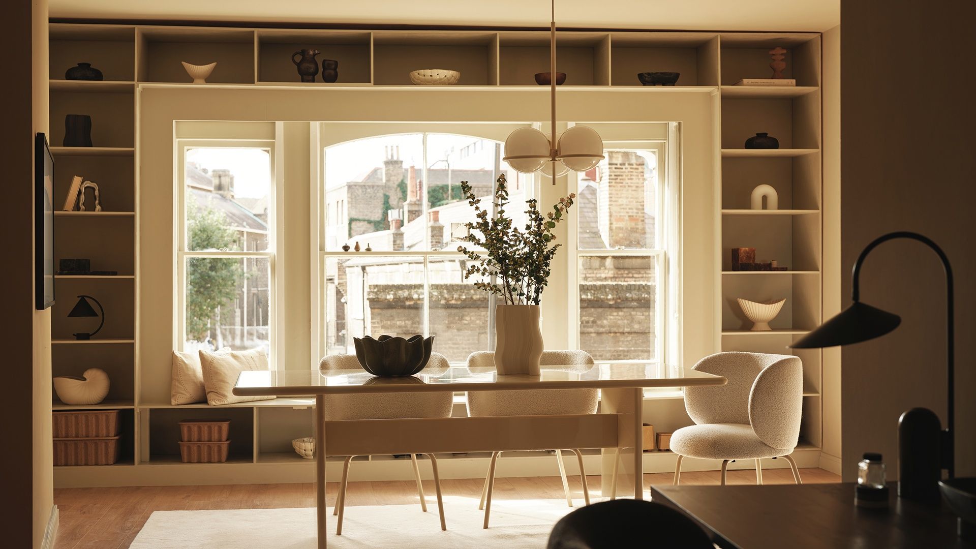 Comedor con mesa de madera, alfombra, sillas de comedor tapizadas, estantería alrededor de un gran ventanal, lámpara de techo, jarrón con ramas verdes