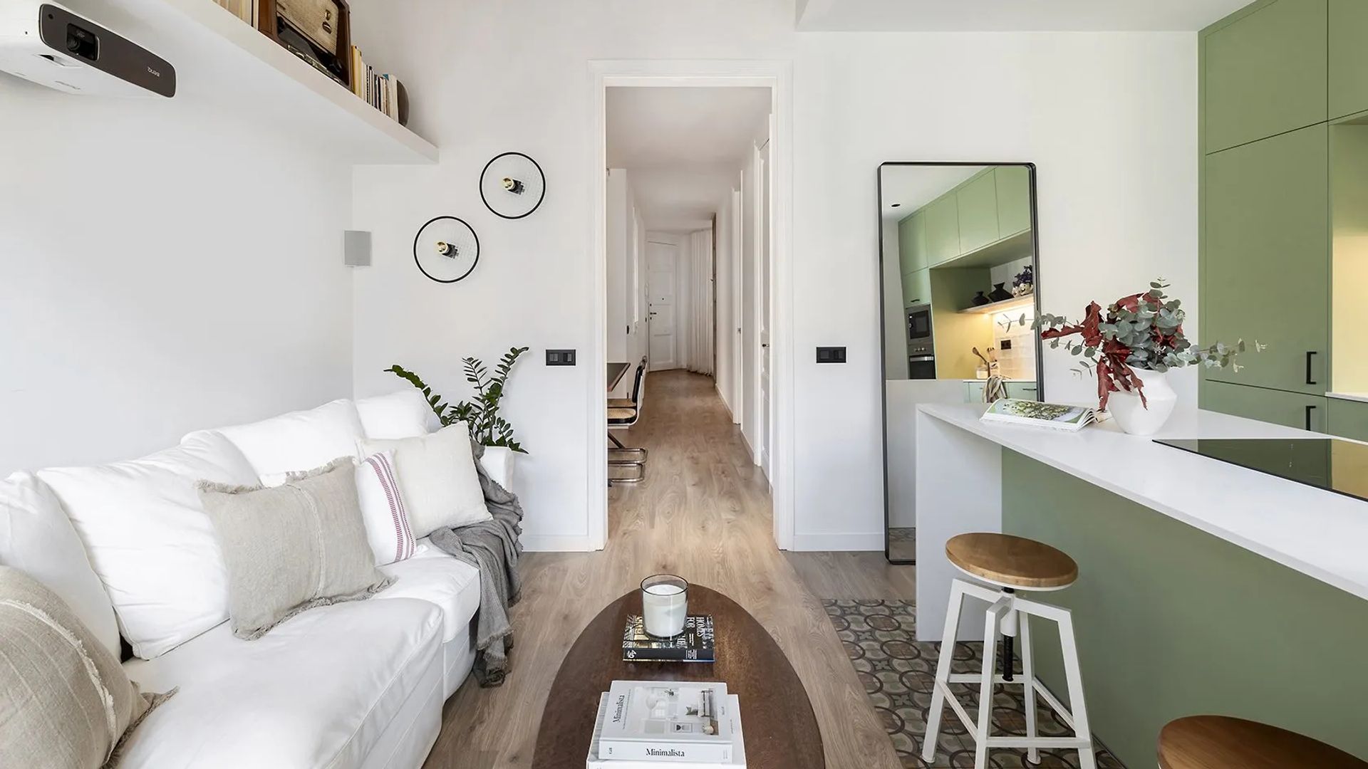 Cocina abierta al salón, con isla, espejo, sofá blanco, vigas en el techo, balda sobre el sofá, muebles de cocina verdes 