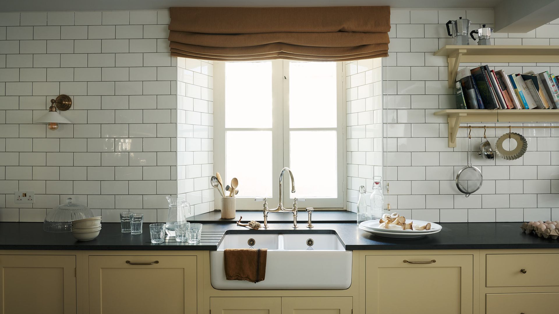 Cocina con fregadero y grifería antigua, encimera negra, pared de azulejos blancos brillantes, muebles color vainilla, estor marrón, baldas con libros 