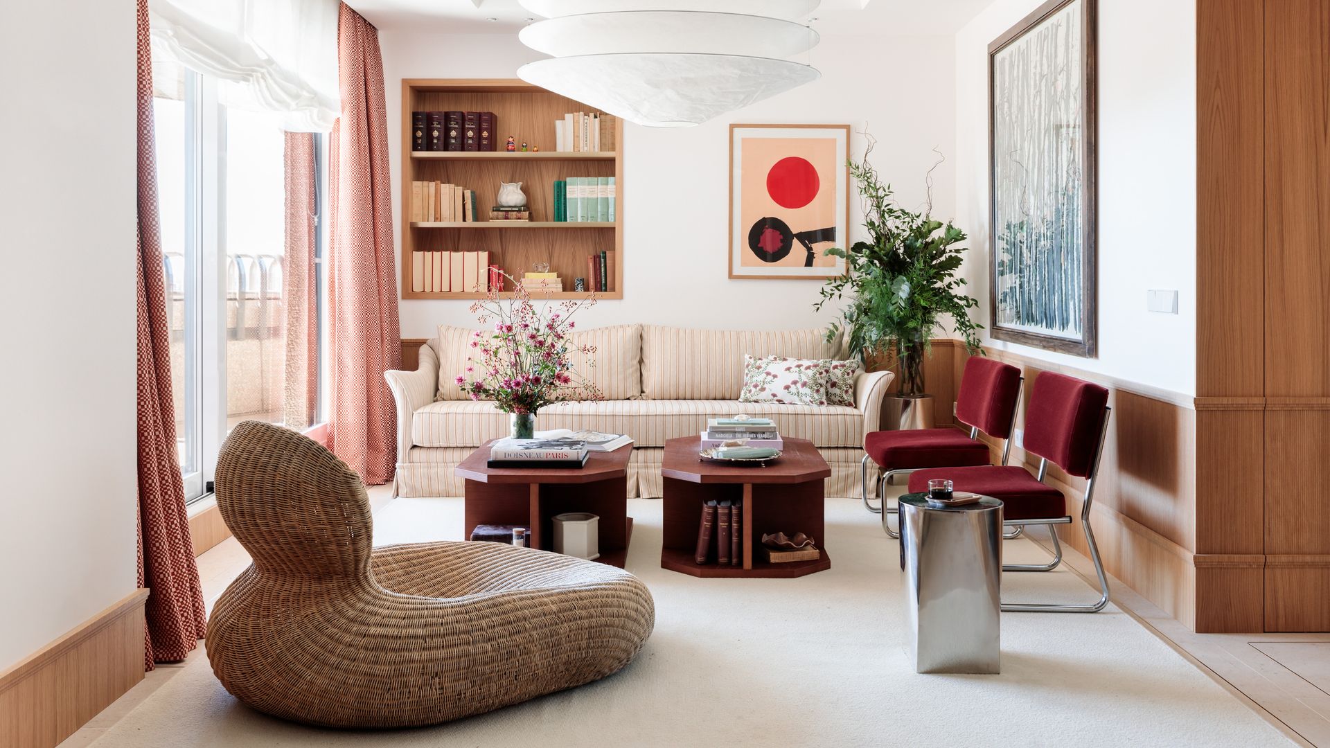 Salón con hornacina en la pared forrada de madera y con baldas, mesas de centro de madera oscura, sillas tapizadas en color burdeos, lámpara de techo XL, cuadros en la pared, sofá beige con tela a rayas, butaca de fibras vegetales