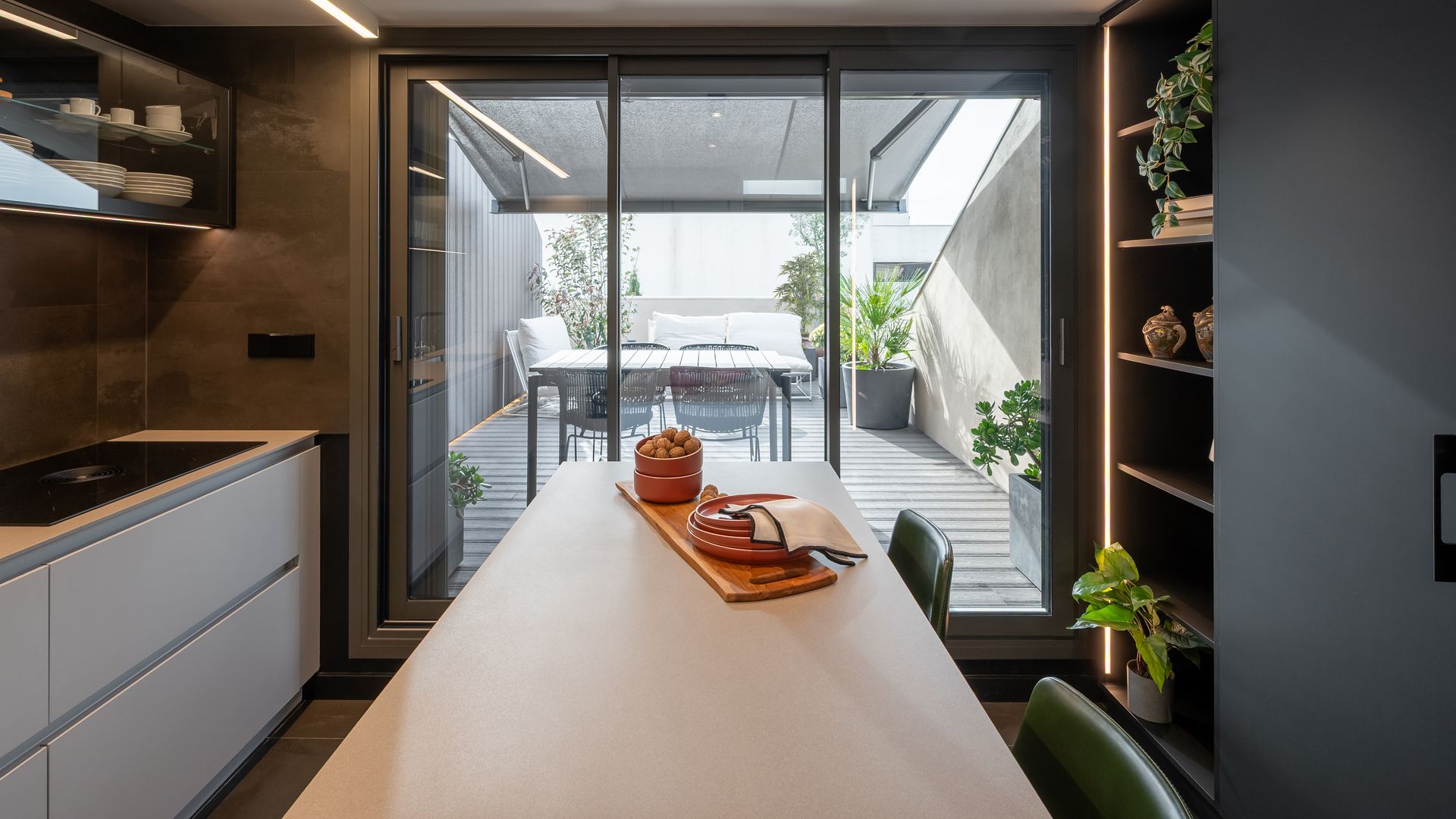 Cocina con salida a la terraza con suelo de madera, mesa de comedor, plantas y toldo negro 