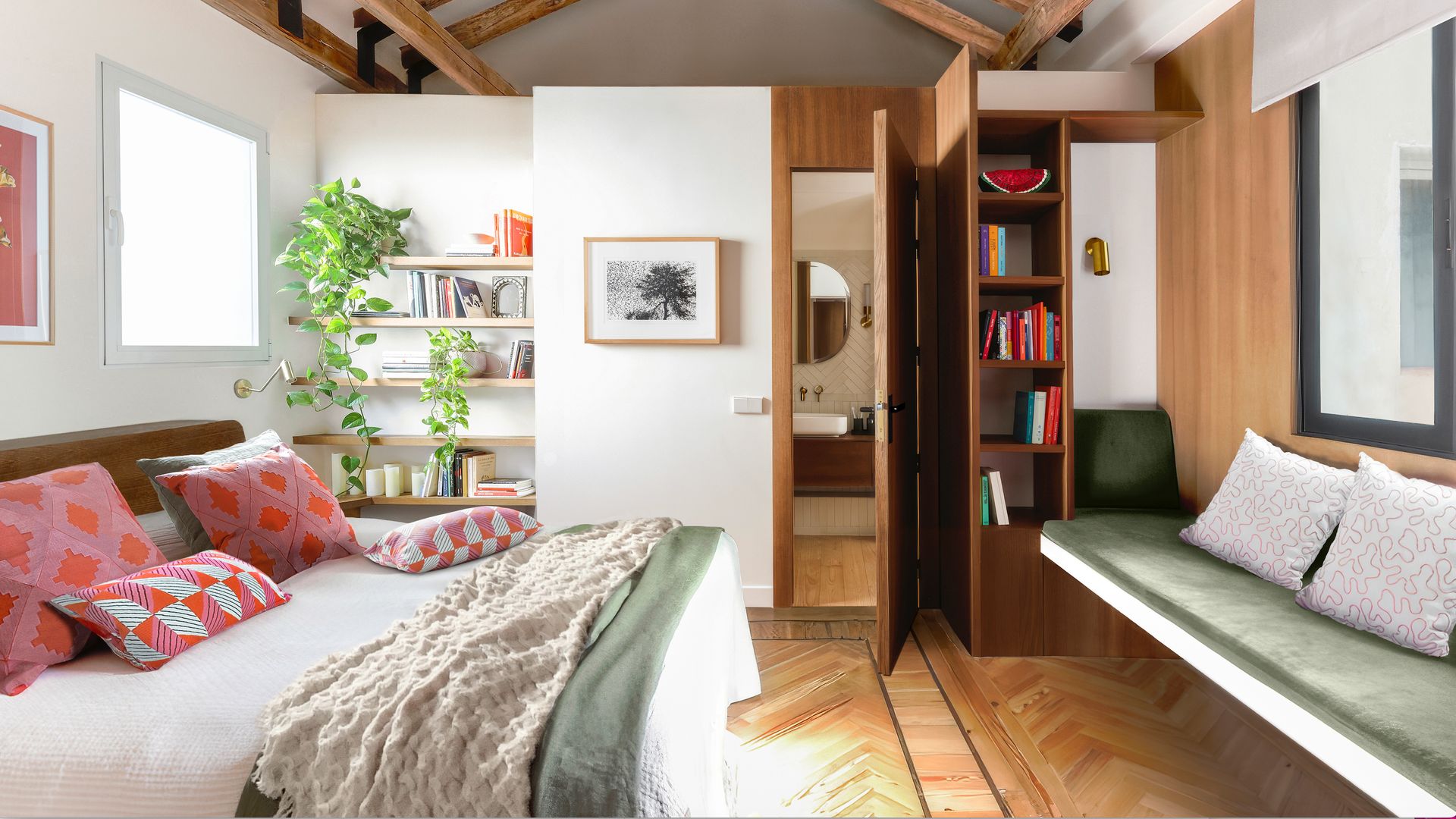 Dormitorio con vigas de madera en el techo, suelo de madera en espiga, baldas blancas, estructura de madera con librería y banco, plantas, cojines en rosa y rojo