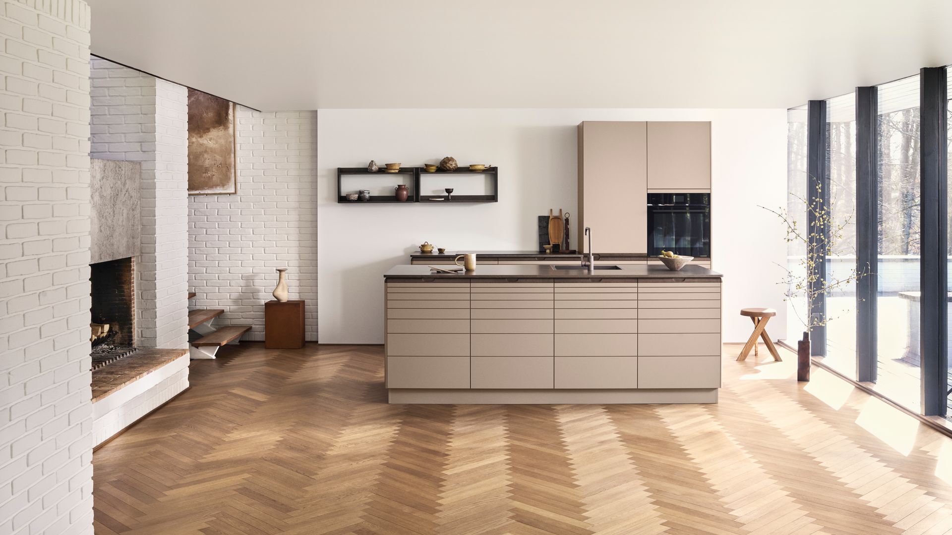 Cocina con isla en tonos beige, suelo de madera, ventanales con salida a la terraza, encimera negra, pared de ladrillo blanco
