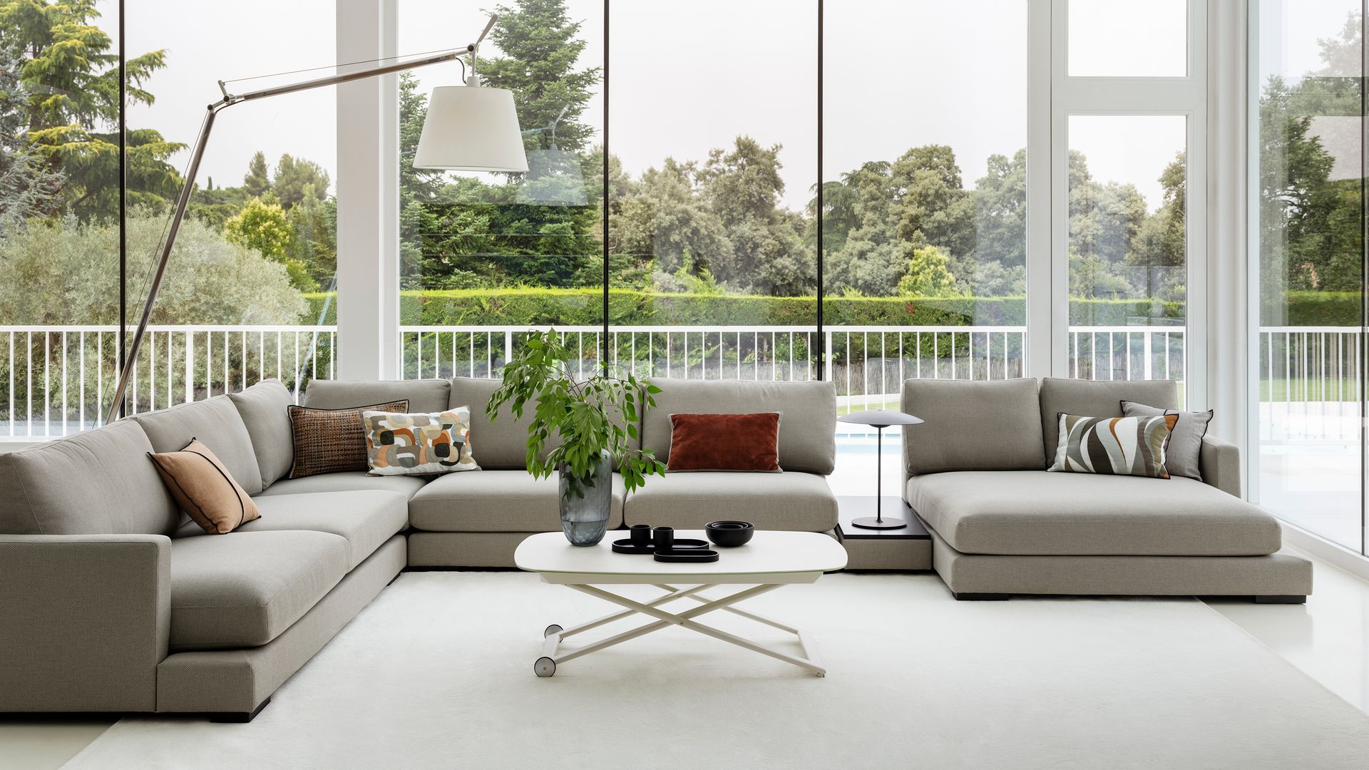 Salón con grandes ventanales con salida al jardín, sofá gris, mesa de centro blanca, alfombra blanca y lámpara de pie