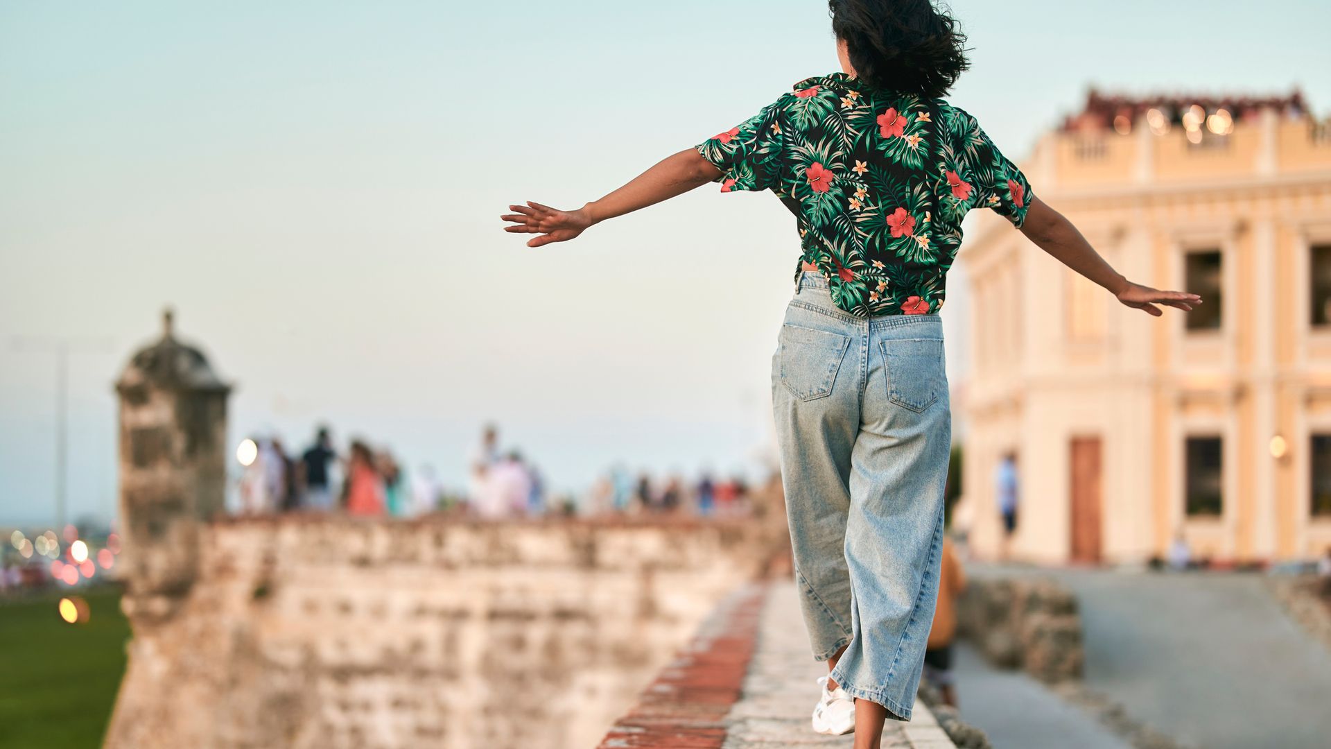 Mujer caminando por encima de una de las murallas de Cartagena, en Colombia. Lleva pantalón vaquero, deportivas blancas y una camisa floreada. Está de espaldas con los brazos abiertos para mantener el equilibrio.