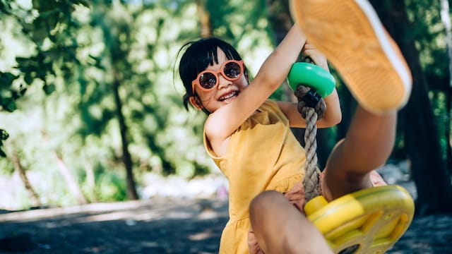 Niña columpiándose en un columpio de colores de árbol, con un vestido amarillo y gafas de sol naranjas. Está disfrutando muy sonriente.