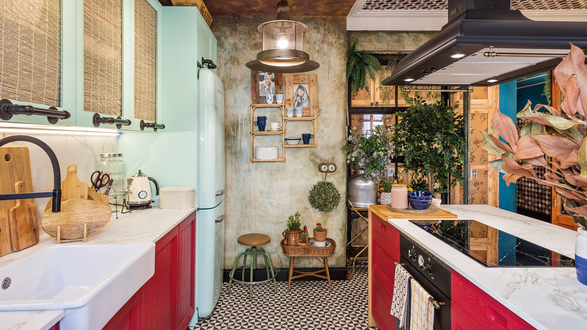 Cocina con muebles rojos, suelo de damero blanco y negro, armarios superiores verde con puertas de fibras vegetales, adornos en las paredes, taburete con plantas