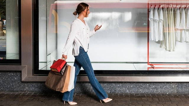 Mujer joven mirando el teléfono móvil mientras camina con bolsas de compras por delante del escaparate de El Corte Inglés