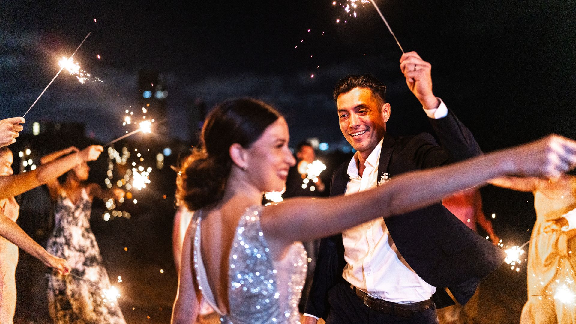 Novios en la fiesta de su boda con bengalas, él con traje y ella con vestido de lentejuelas, sonrientes y rodeados de amigos