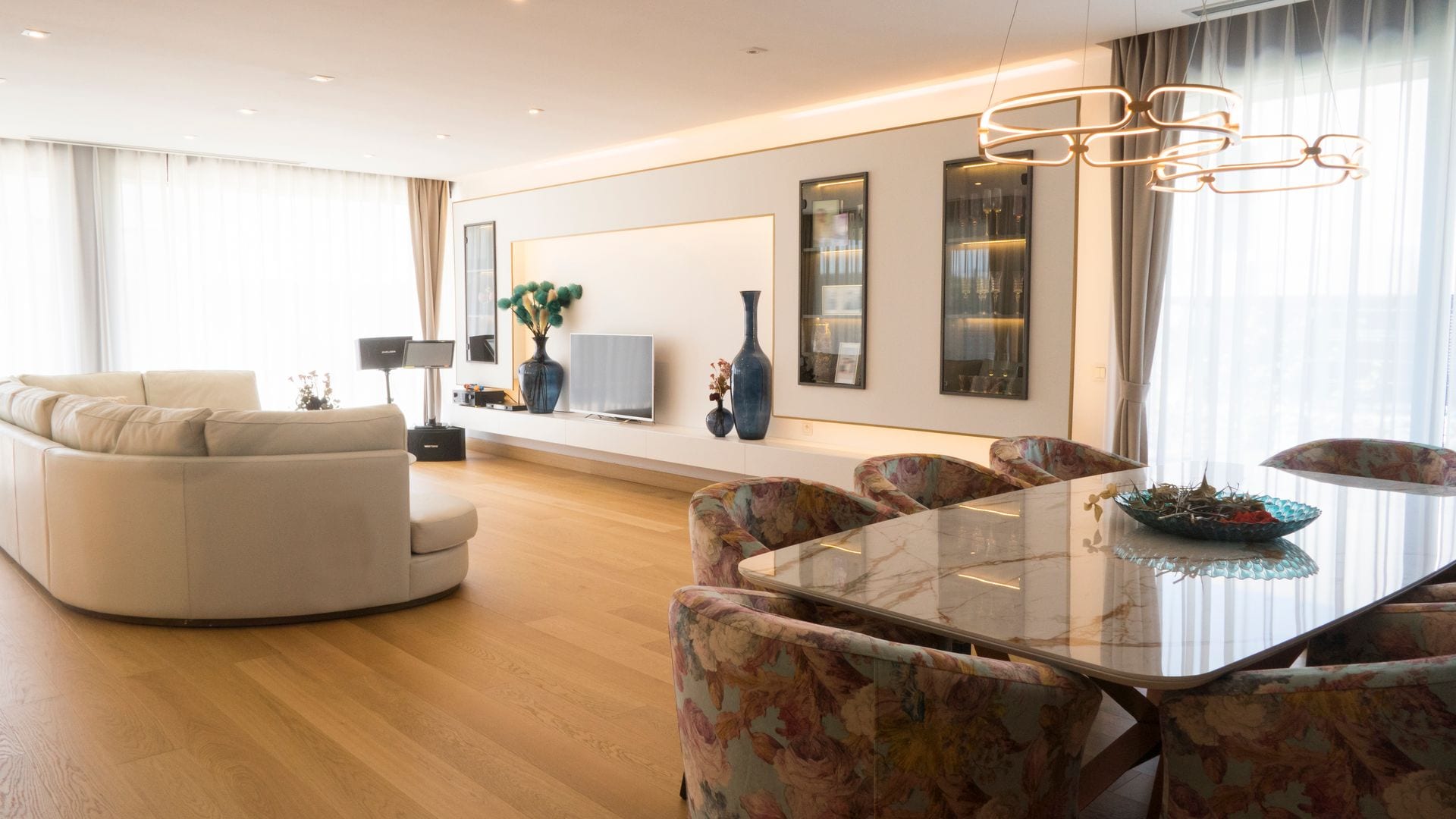Salón comedor con mesa de comedor, sillas de comedor tapizadas, lámpara de techo, sofá blanco, suelo de madera, mueble de televisión a medida con vitrinas integradas 