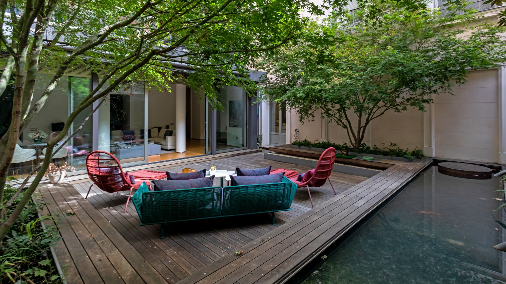 Terraza urbana con suelo de madera, estanque, plantas, muebles de color rojo y verde, grandes árboles 