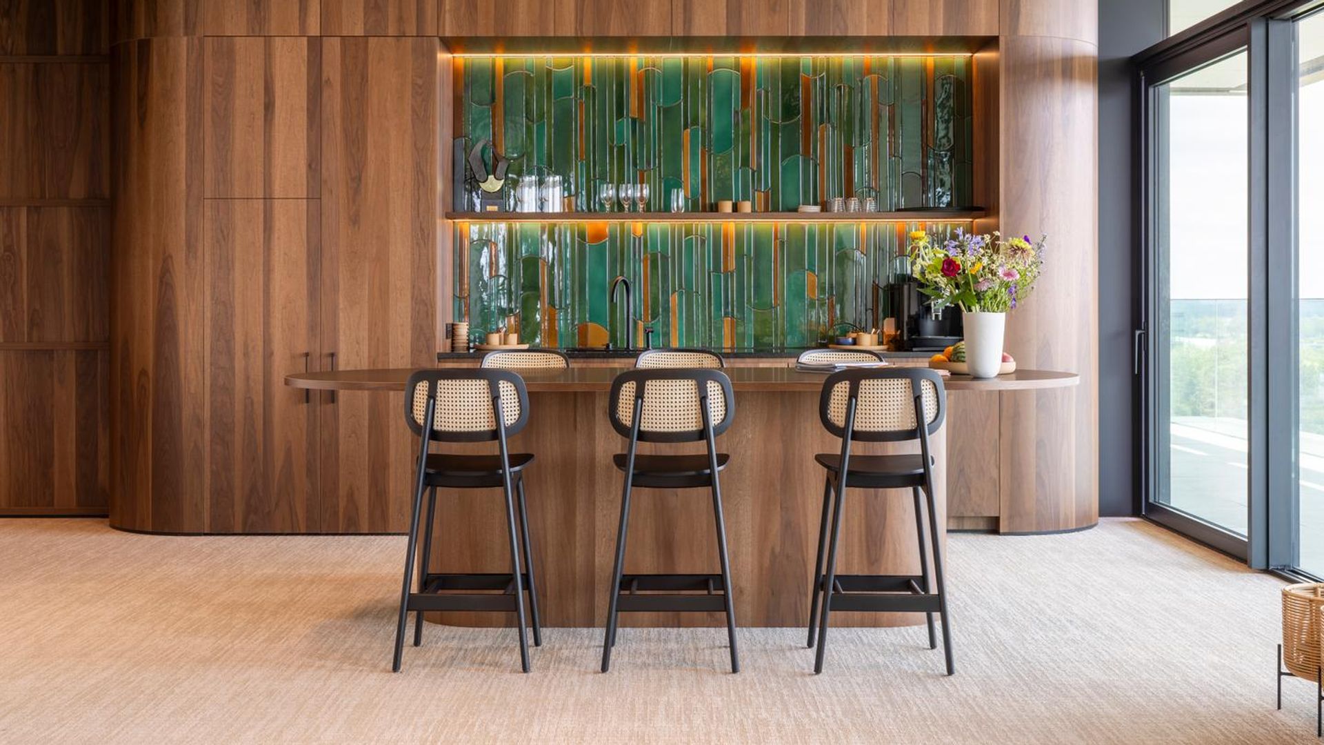 Barra de bar en casa con taburetes de rejilla, isla a modo de madera, ventanal, mueble de madera abierto con baldas con copas y botellas