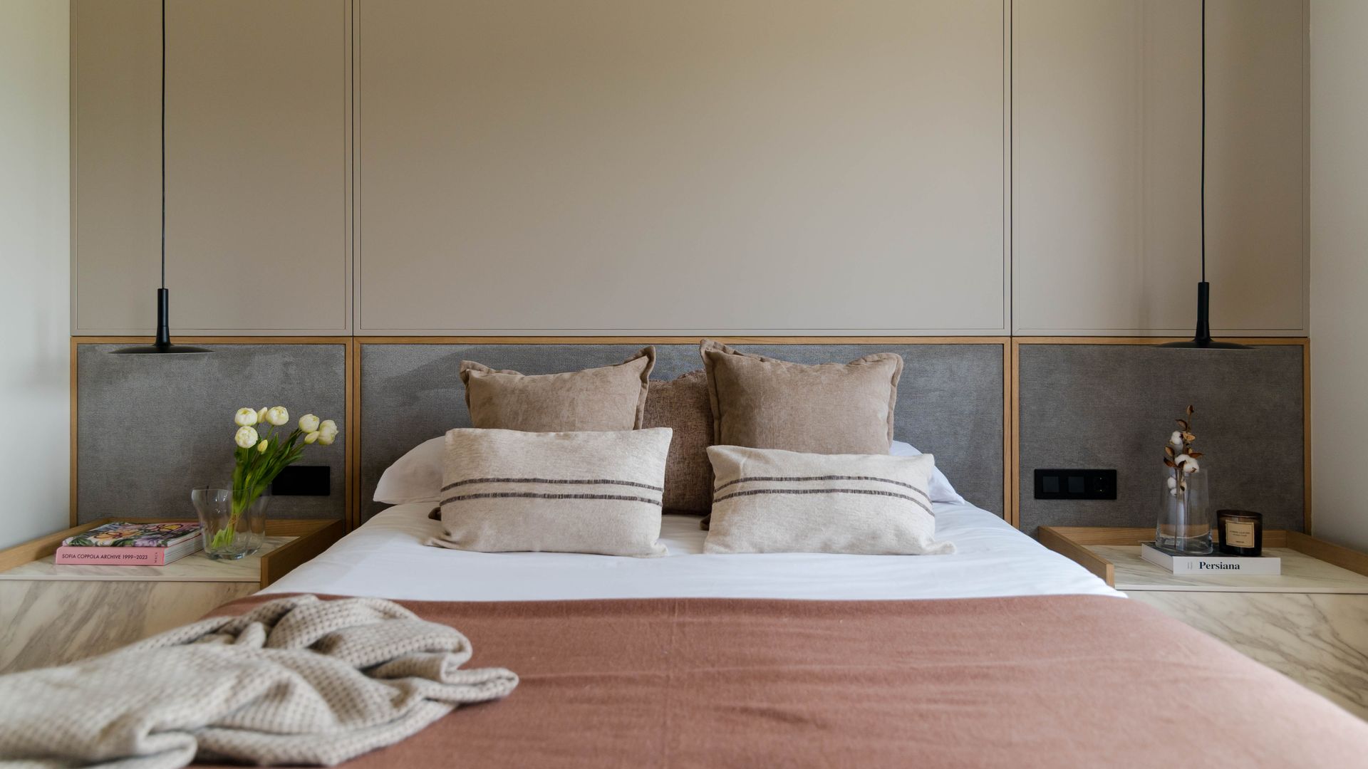 Dormitorio con pared del cabecero tapizado, apliques a los lados de la cama, ropa de cama marrón, cojines
