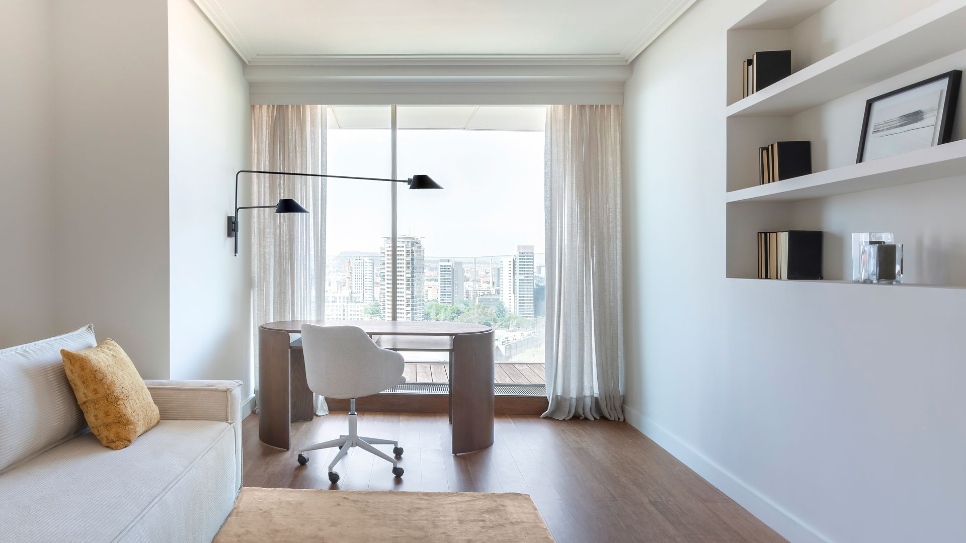 Despacho con mesa de escritorio, ventana, sofá blanco, alfombra, librería de obra, cortinas blancas, suelo de madera 