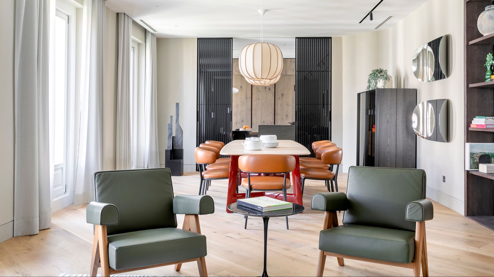 Salón comedor con butacas verdes, sillas de comedor de cuero, lámparas de techo sobre la mesa, cortinas, suelo de madera 
