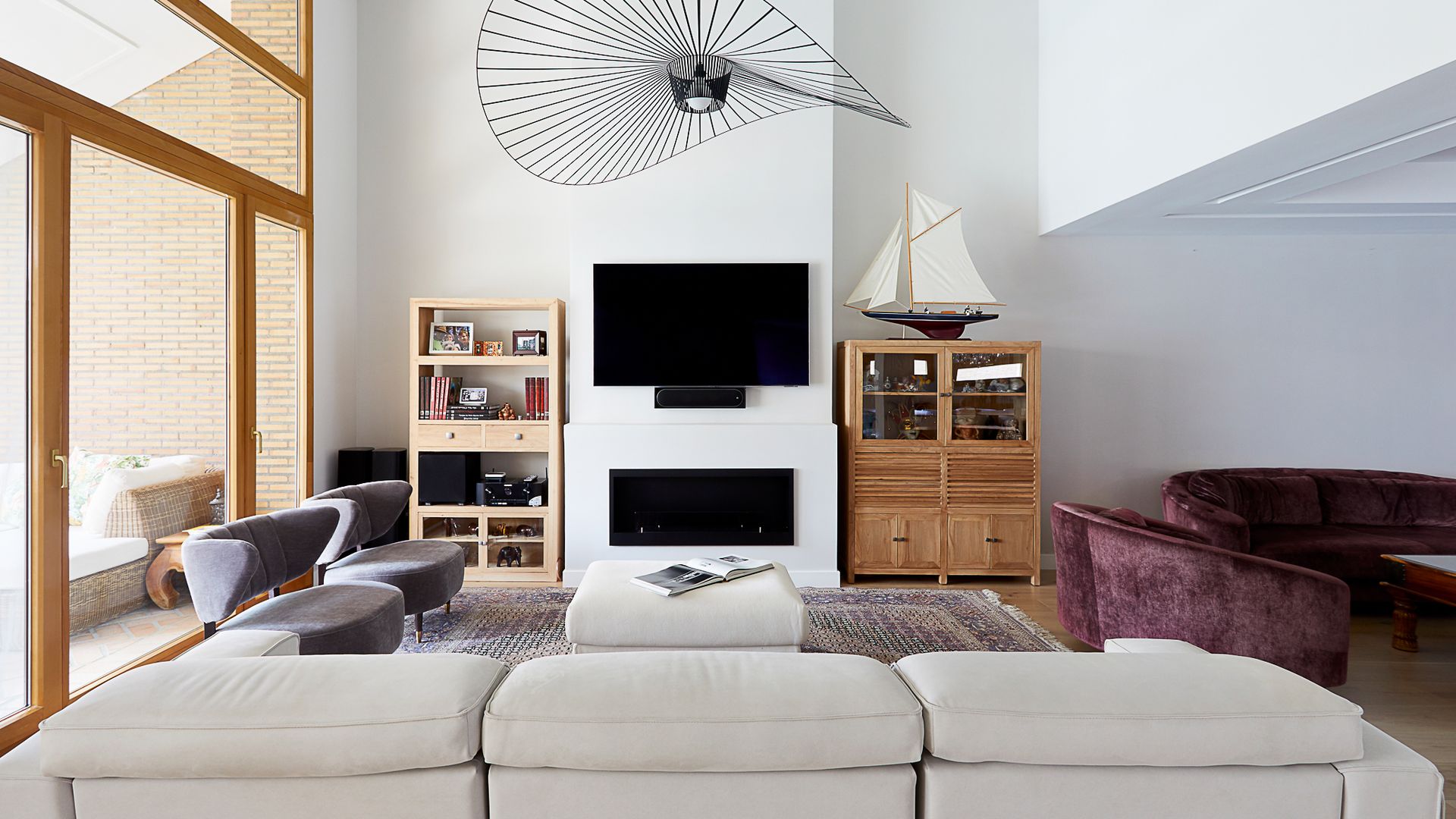 Salón con chimenea, lámpara de techo Vértigo, sofá beige, butacas grises, aparador alto de madera oscura, estantería de madera clara, televisión colgada en la pared 
