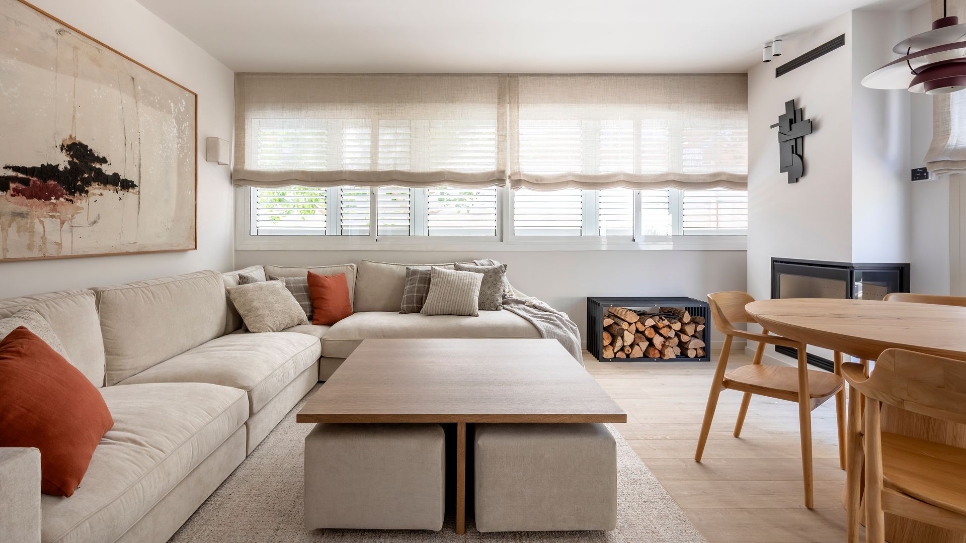 Salón comedor con estores, sofá modular de color beige, mesa de centro de madera con pufs debajo, cuadro en la pared del sofá, chimenea, alfombra