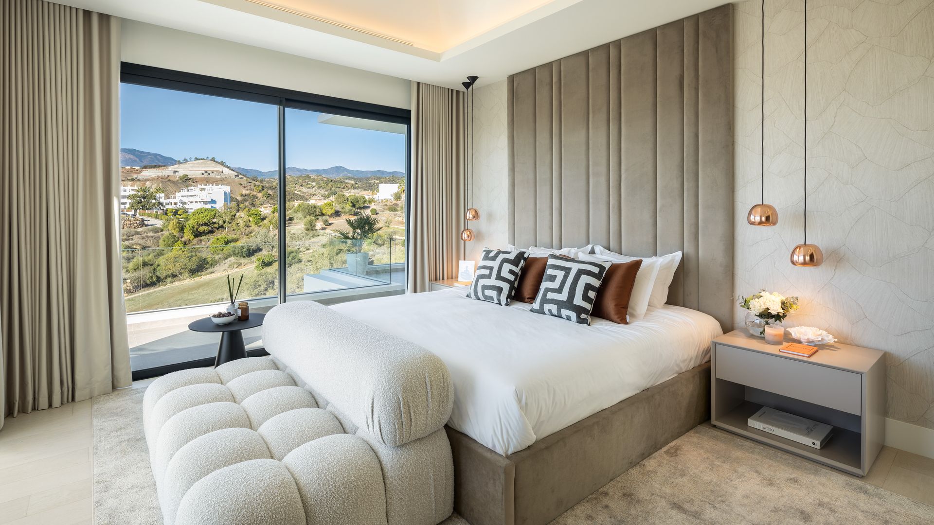Dormitorio con cabecero alto tapizado, cama tapizada, mesilla beige, lámparas de techo en color cobre, sofá blanco a los pies de la cama, salida a terraza, techo retroiluminado