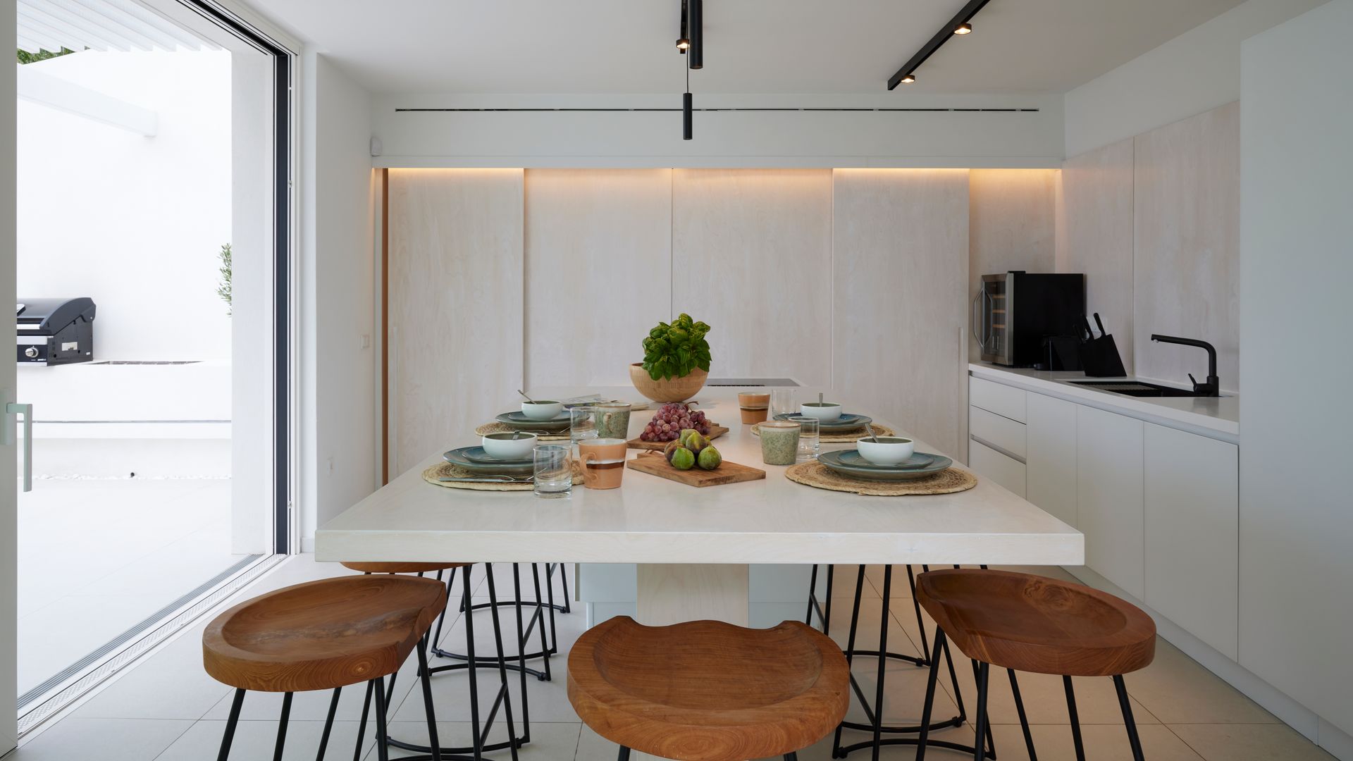 Cocina blanca con armario, luz en el techo, isla central con zona de comedor con taburetes de madera, luz perimetral en el techo e hileras de focos negros