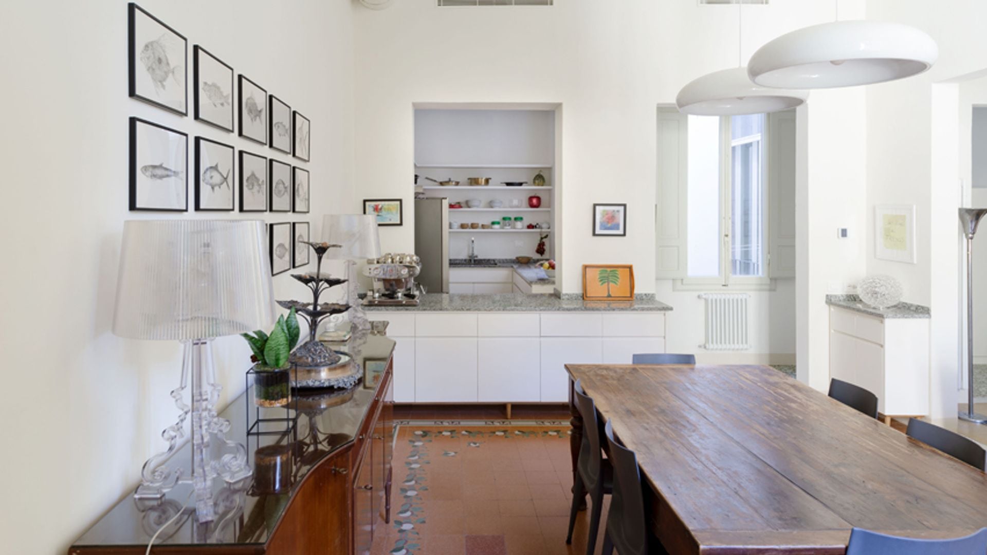 Comedor con suelo de terrazo marrón, mesa de madera antigua, aparador, cuadro en la pared, lámparas de techo blancas, abertura que comunica con la cocina